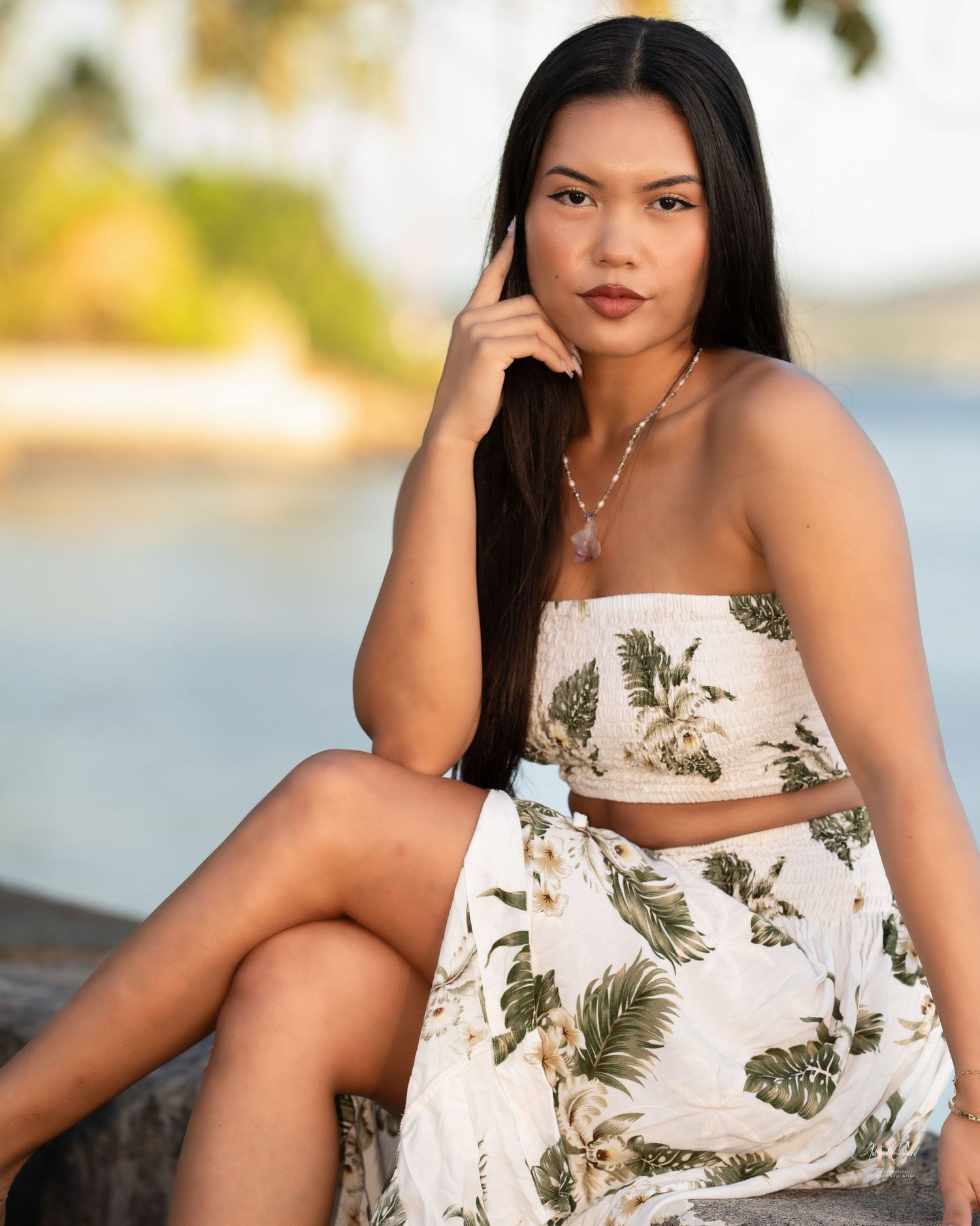 A person in a tropical print two-piece outfit poses on a beach with blurred water and nature in the background.