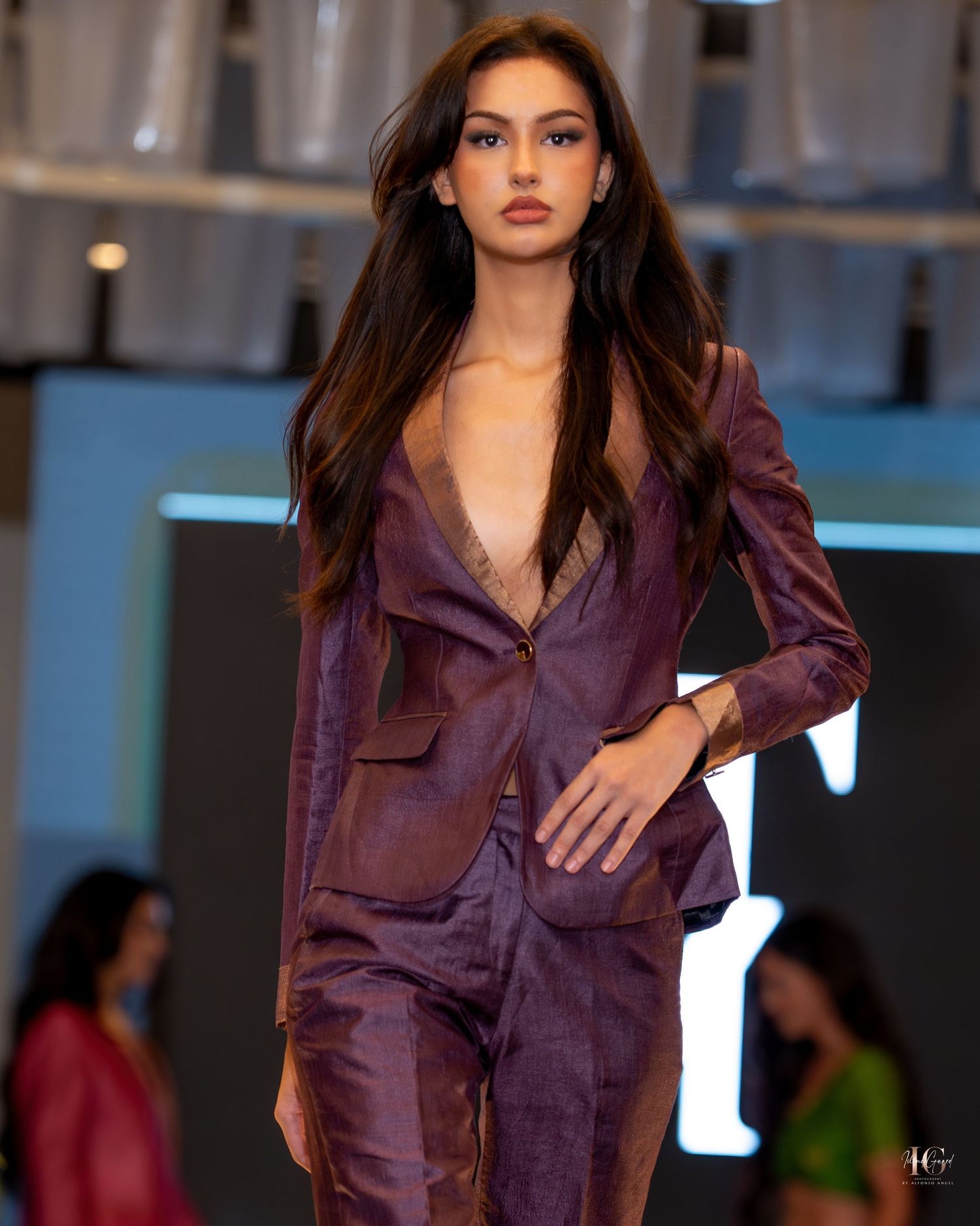 Model walks runway in plum colored satin pantsuit with blazer during fashion show presentation.