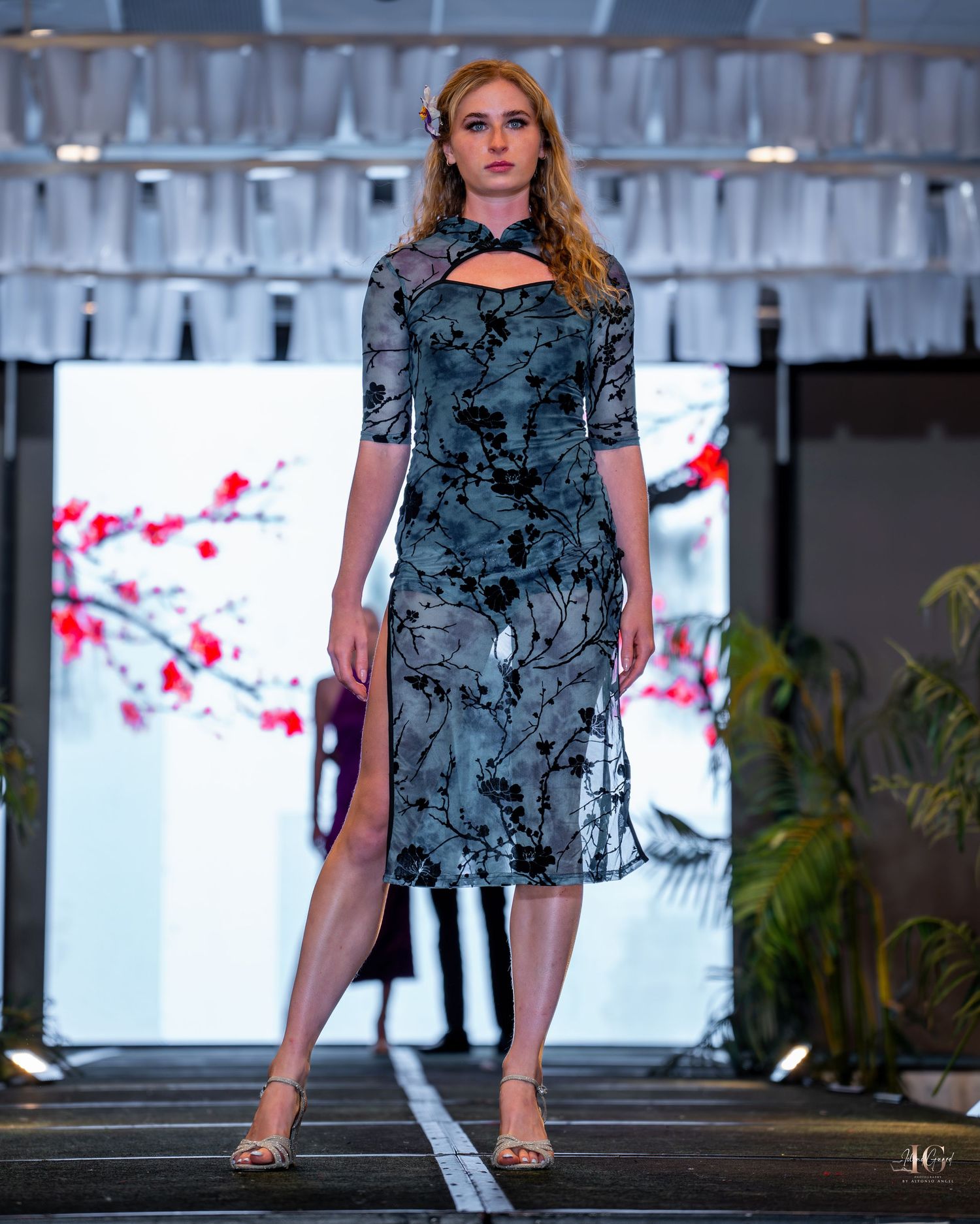 Model walks runway in blue floral print sleeveless top at fashion show event.