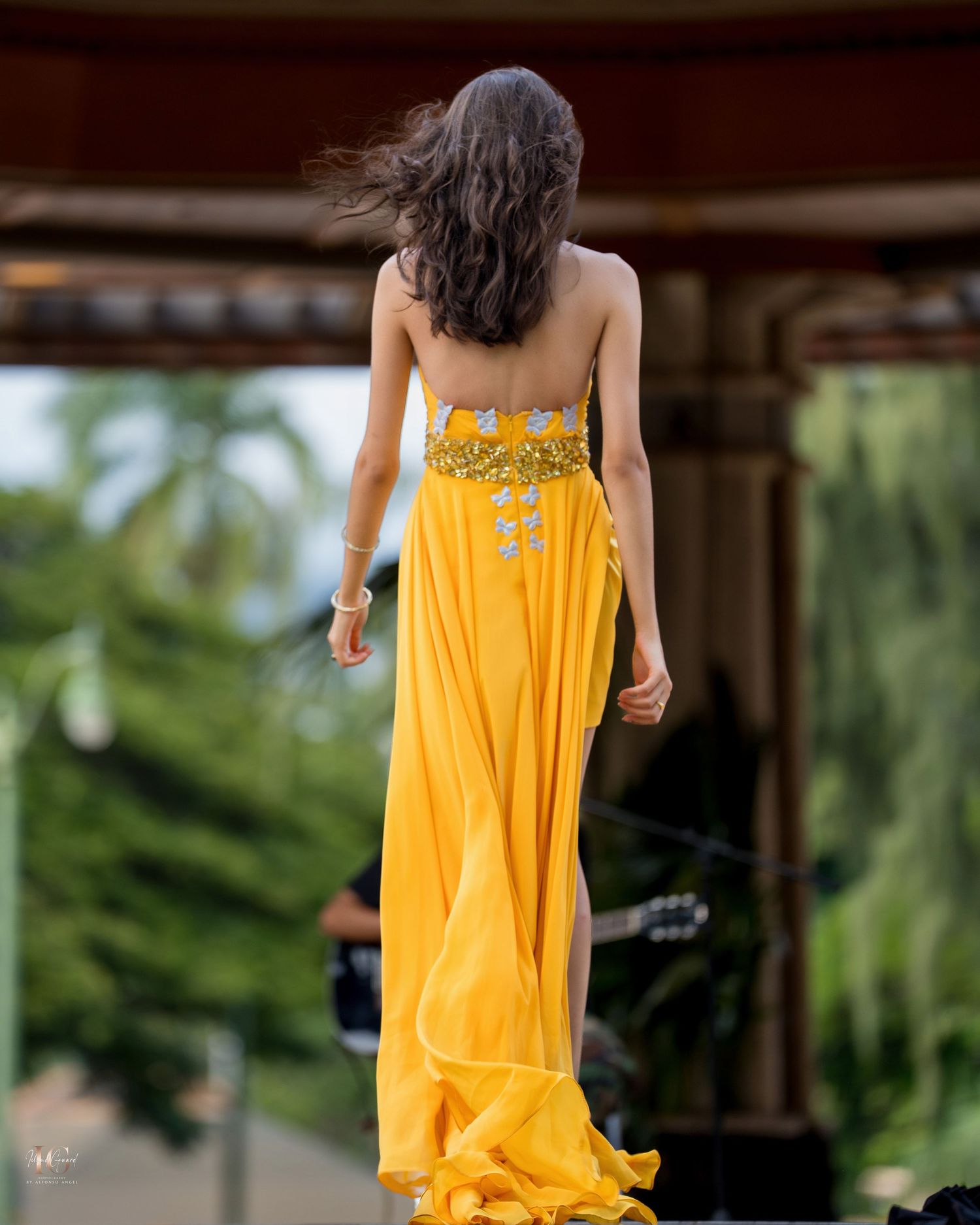 A woman in a flowing yellow sundress walks through a lush garden setting with soft natural lighting.