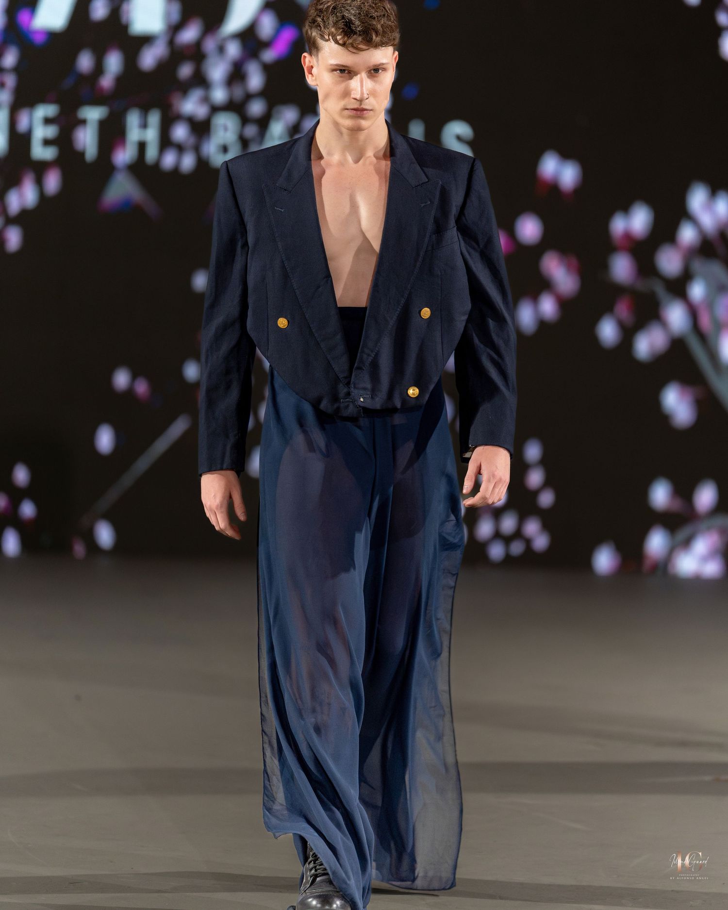 Model walks runway in navy double-breasted blazer during fashion show with purple lighting effects in background.