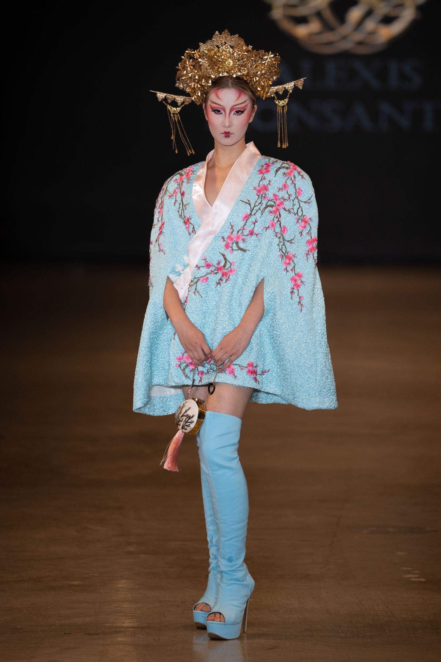 Model walks runway in light blue kimono with cherry blossoms and ornate gold headdress during Asian-inspired fashion show.