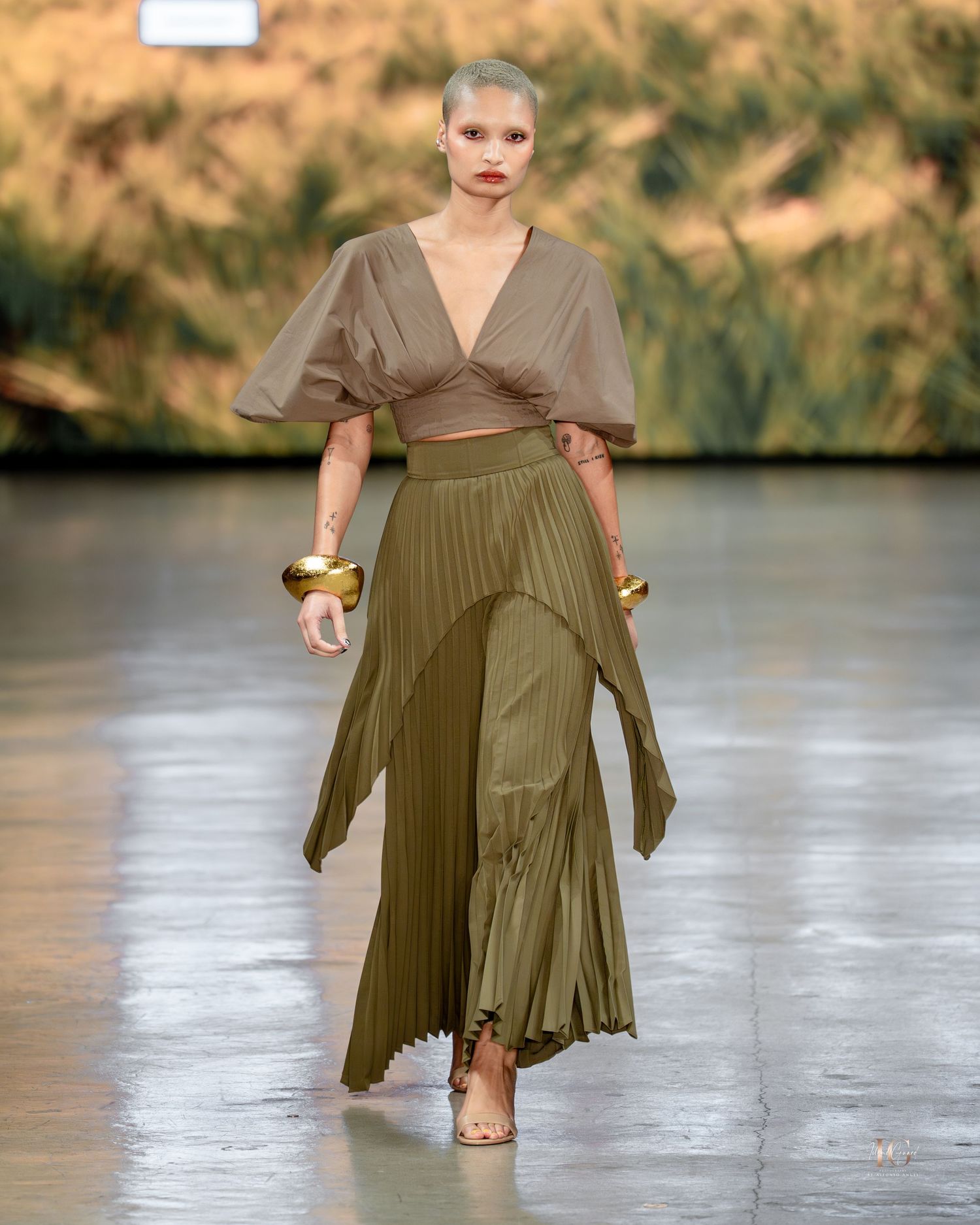 Model walks runway in olive green pleated wrap dress with short sleeves during fashion show.
