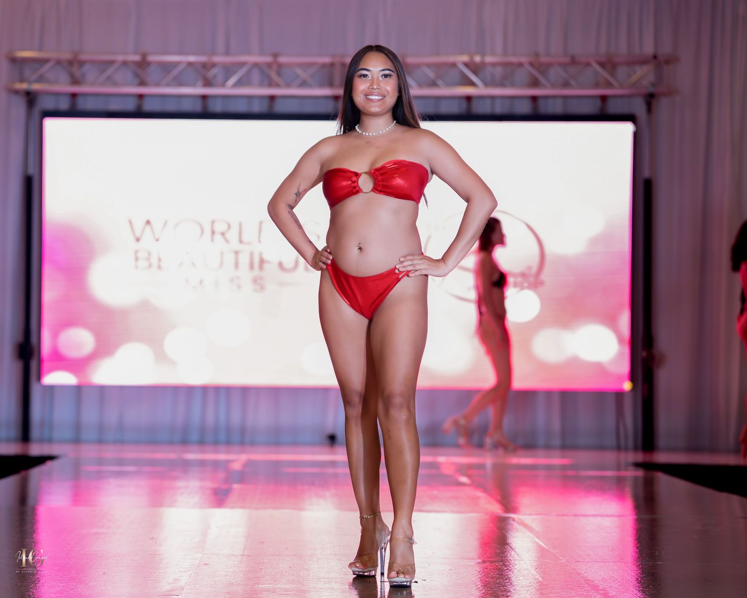 Model walks runway wearing red two-piece swimwear during fashion show with pink lighting and reflective floor.