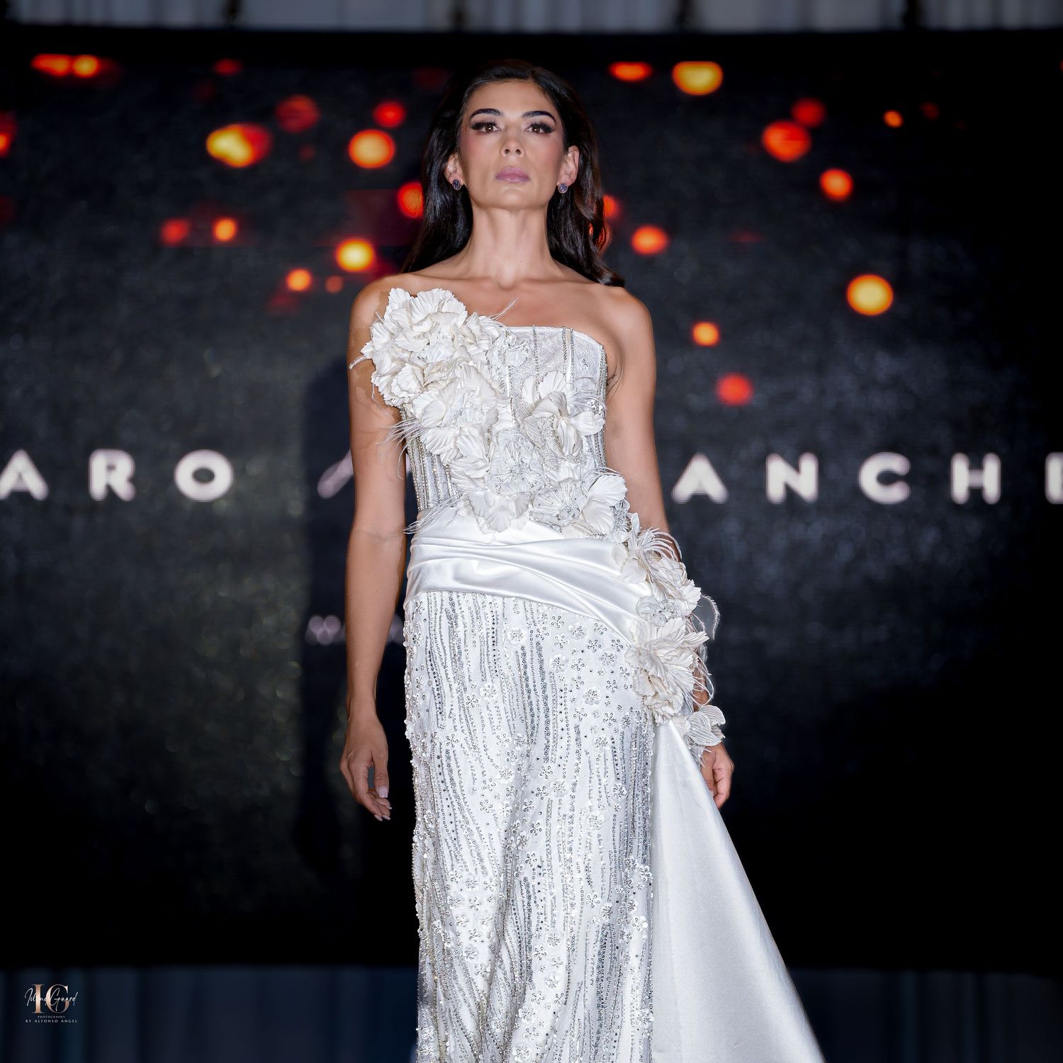 Elegant white dress with floral appliques showcased on catwalk under dramatic lighting.