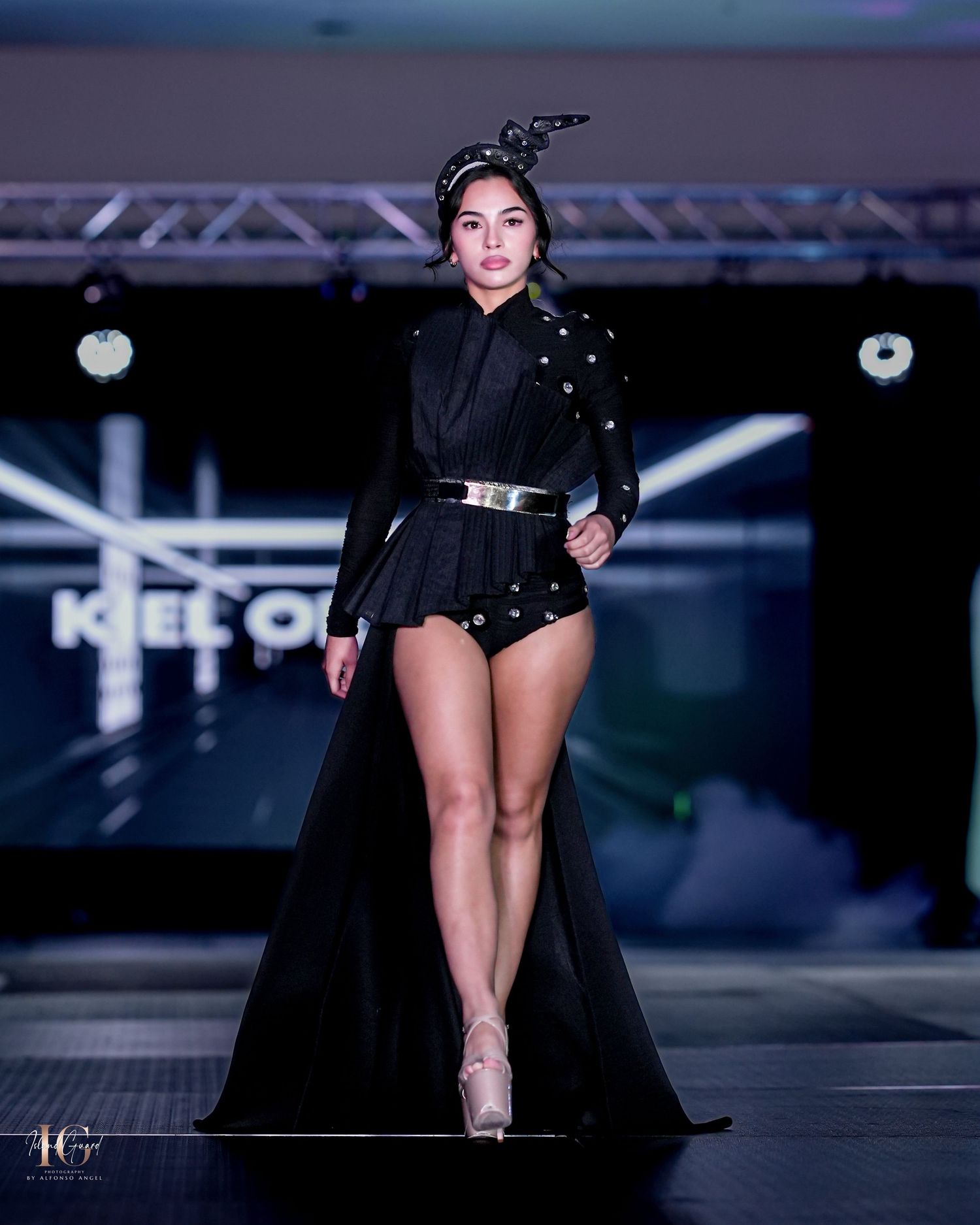 Model showcases avant-garde black high-low dress with belt detail on illuminated catwalk.