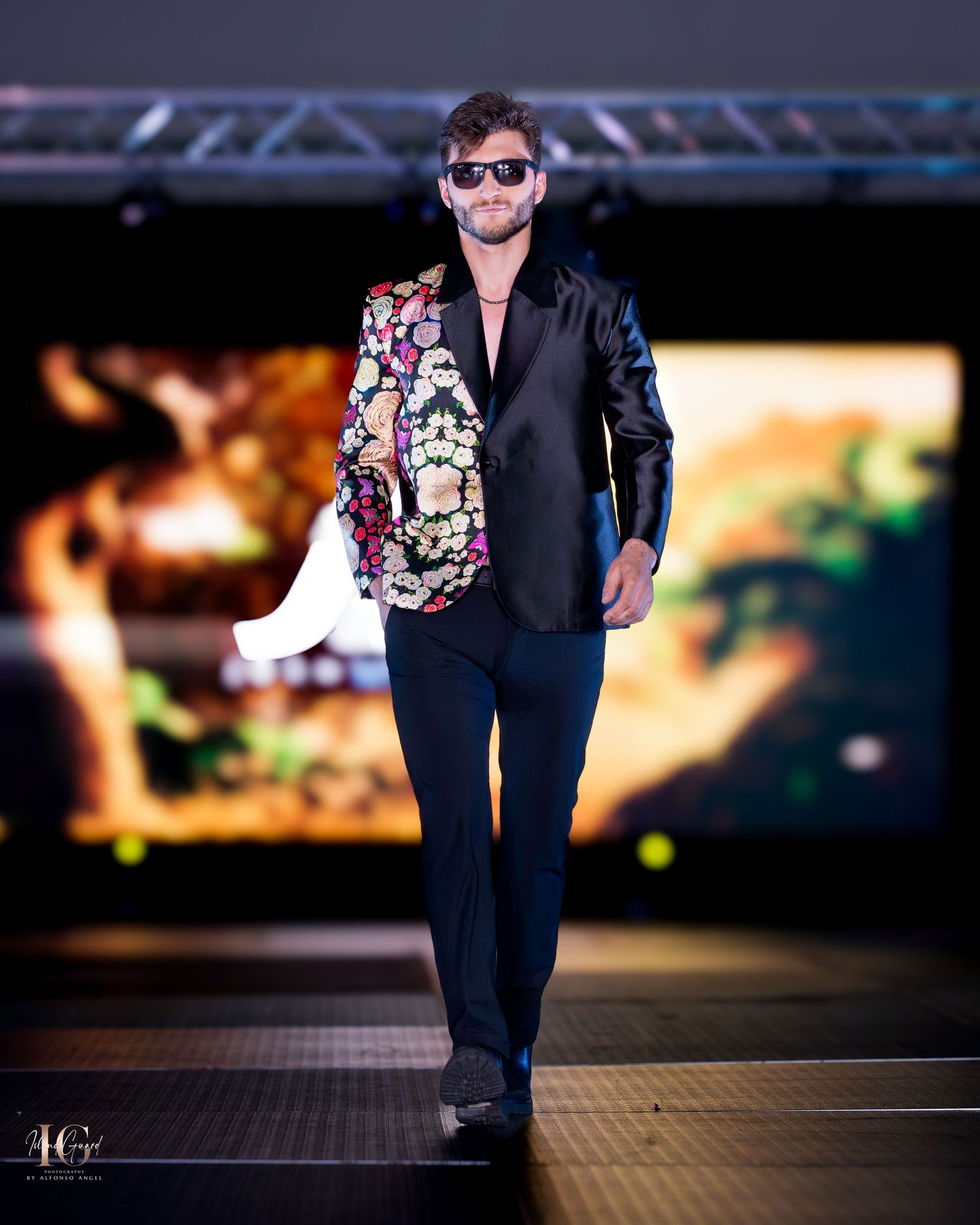 Fashion show model wearing dark blazer with floral print details walks the runway.