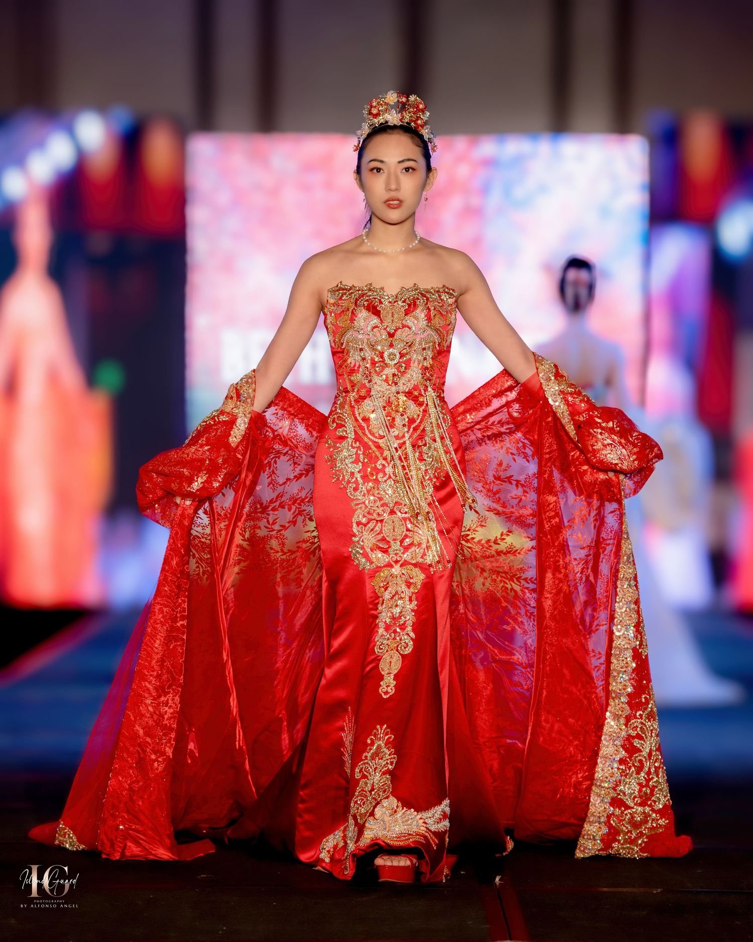 Dramatic red ball gown with gold embellishments showcased on illuminated fashion runway.