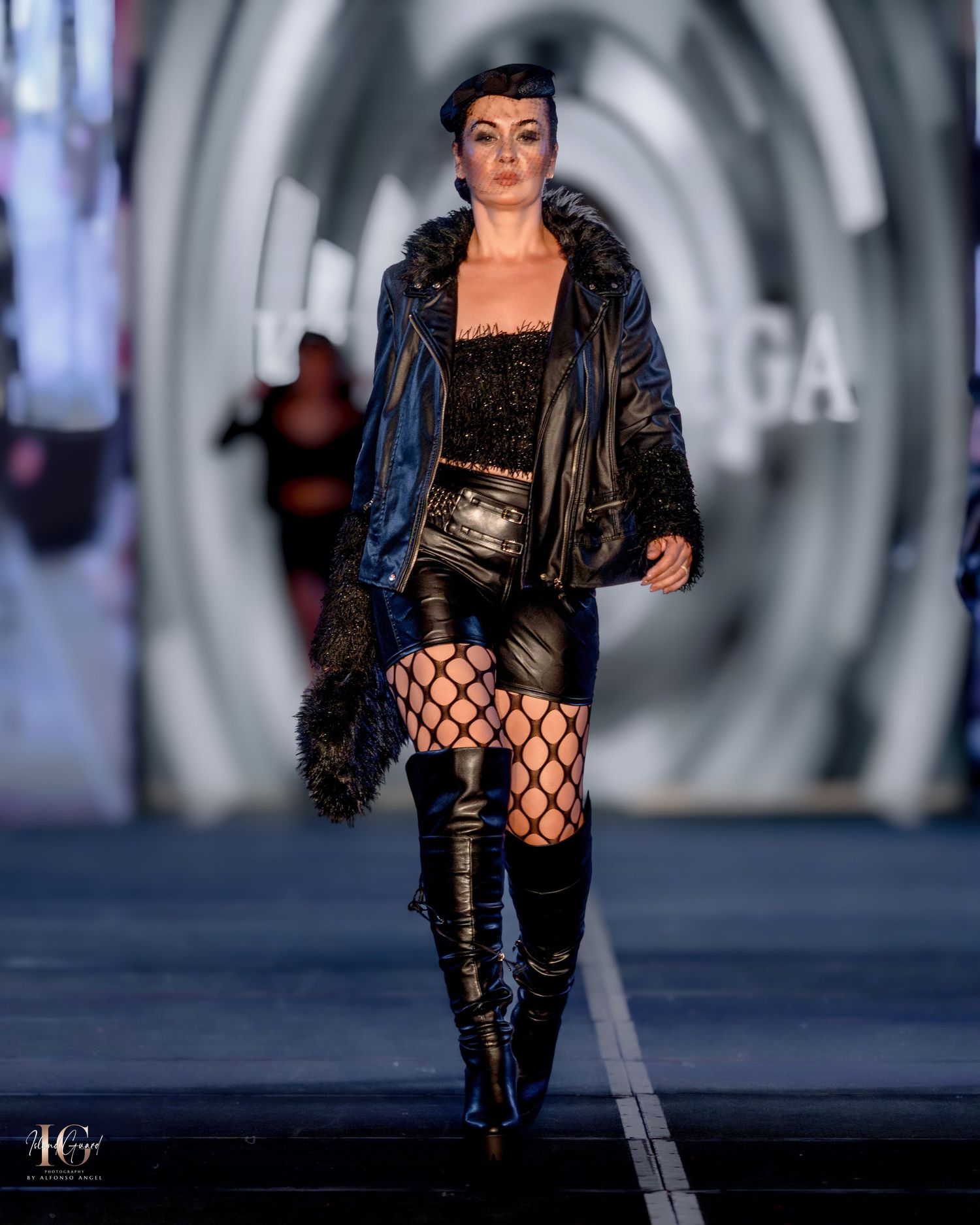 Fashion model wearing leather jacket and fishnet stockings with thigh-high boots on catwalk.