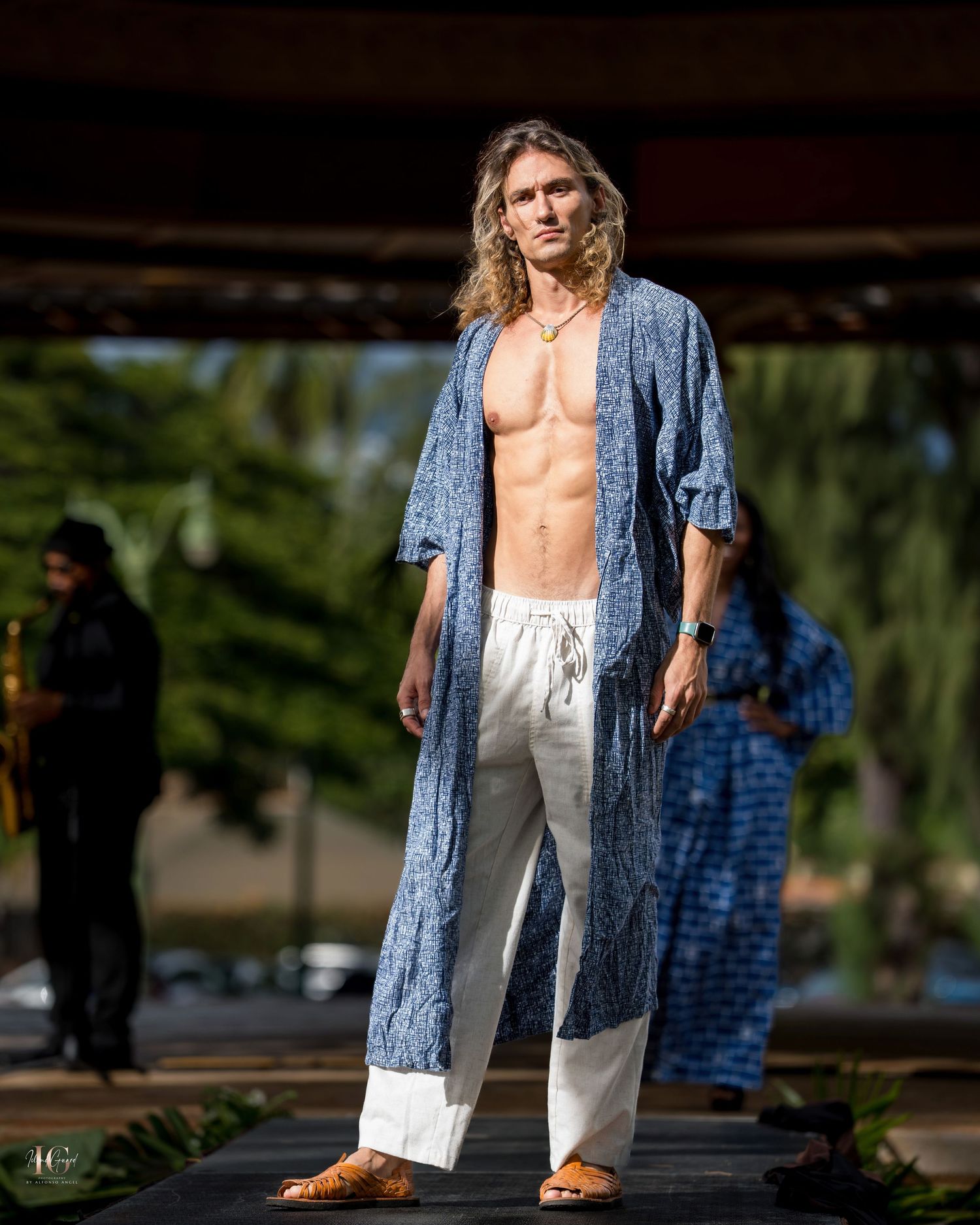 Model wearing unbuttoned denim jacket and white pants poses outdoors in natural sunlight against blurred background.