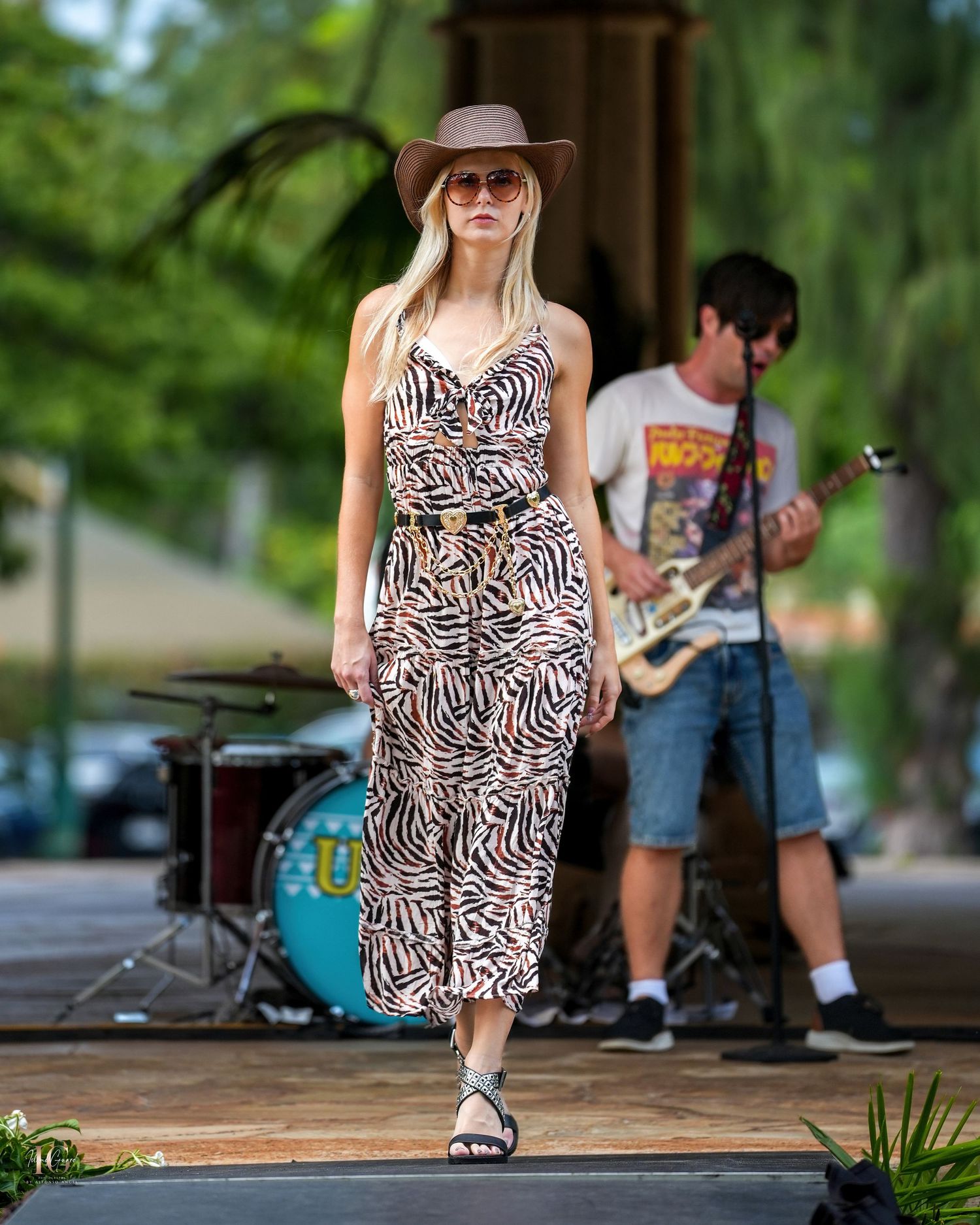 A musician in a zebra print dress and brown hat performs on an outdoor stage while a guitarist plays in the background.