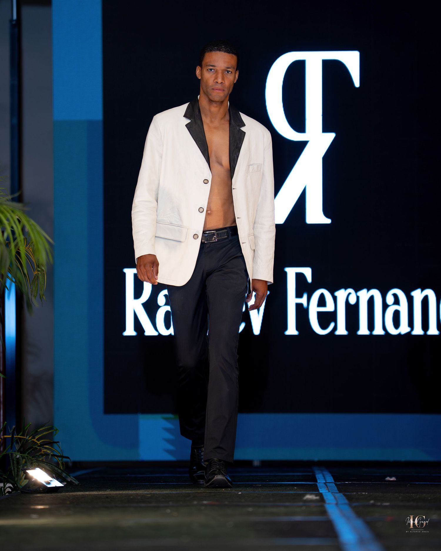 Model walks the runway in a white blazer and black pants during a fashion show with dark blue backdrop.