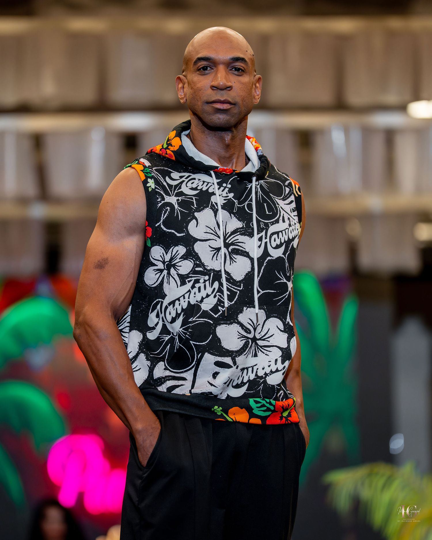 Person wearing a black and white floral print sleeveless shirt performs on stage with colorful lighting in background.