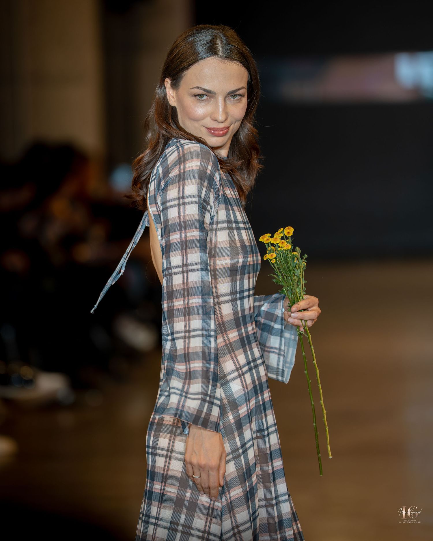 Model walks runway in plaid dress holding yellow flowers at fashion show.