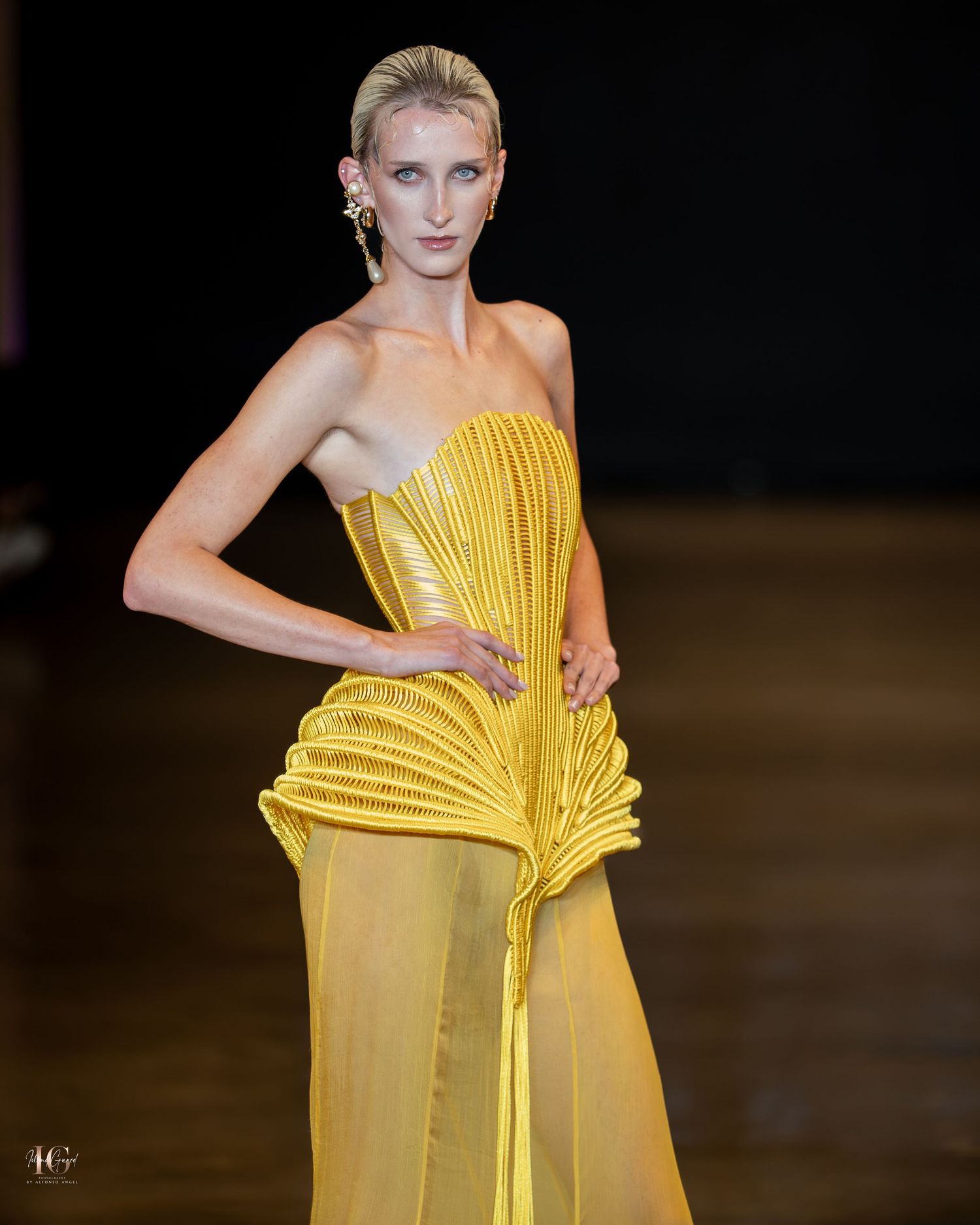 Model showcases a striking yellow pleated evening gown with architectural details on a dark runway.