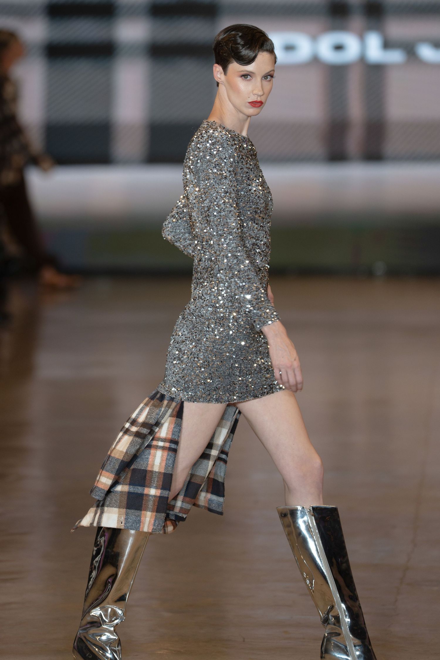 Model walks runway in sparkly silver mini dress with plaid shirt and metallic knee-high boots at fashion show.