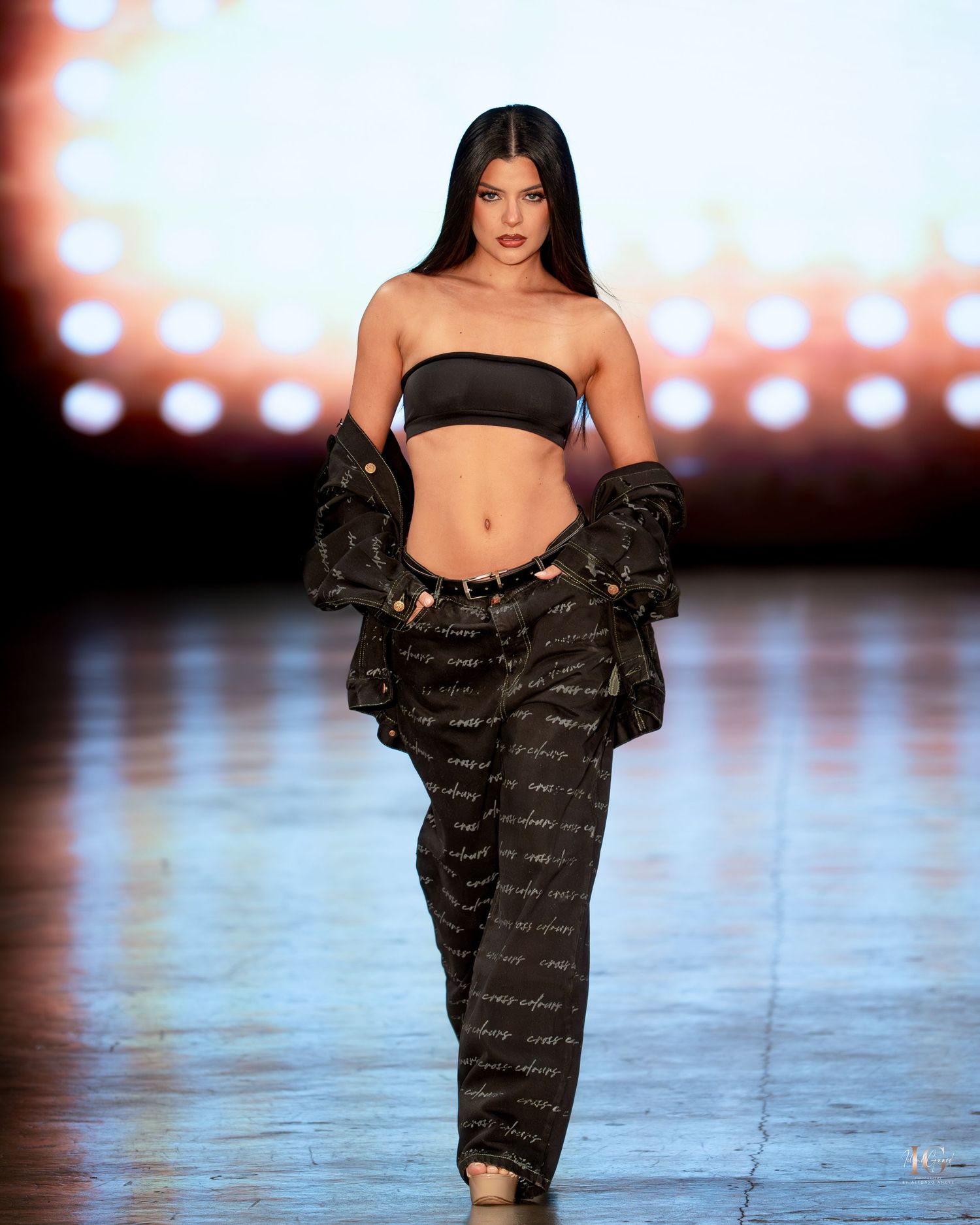 Model walks runway in black bandeau top and oversized printed cargo pants with matching jacket at fashion show.