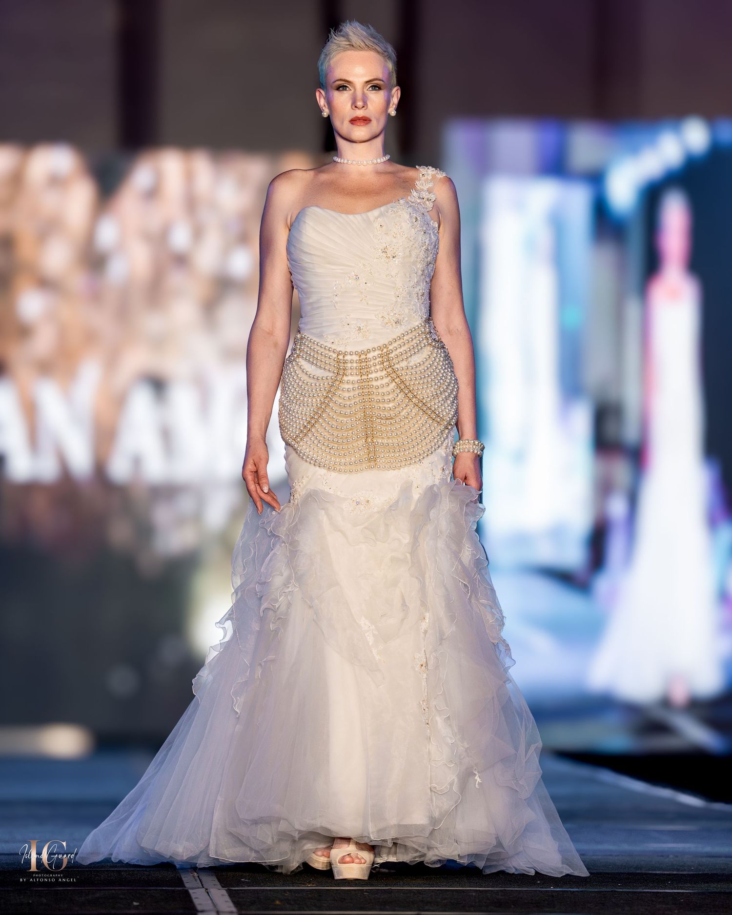 Model walks runway in cream strapless wedding gown with structured bodice and flowing tulle skirt at fashion show.