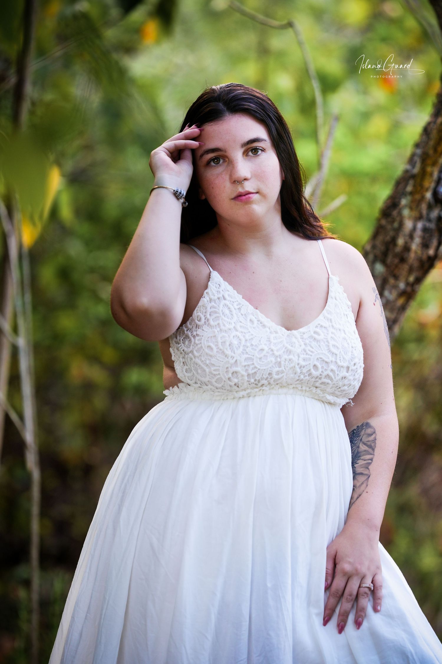 A flowing white lace maternity gown photographed in a natural outdoor setting with soft bokeh and greenery in the background.