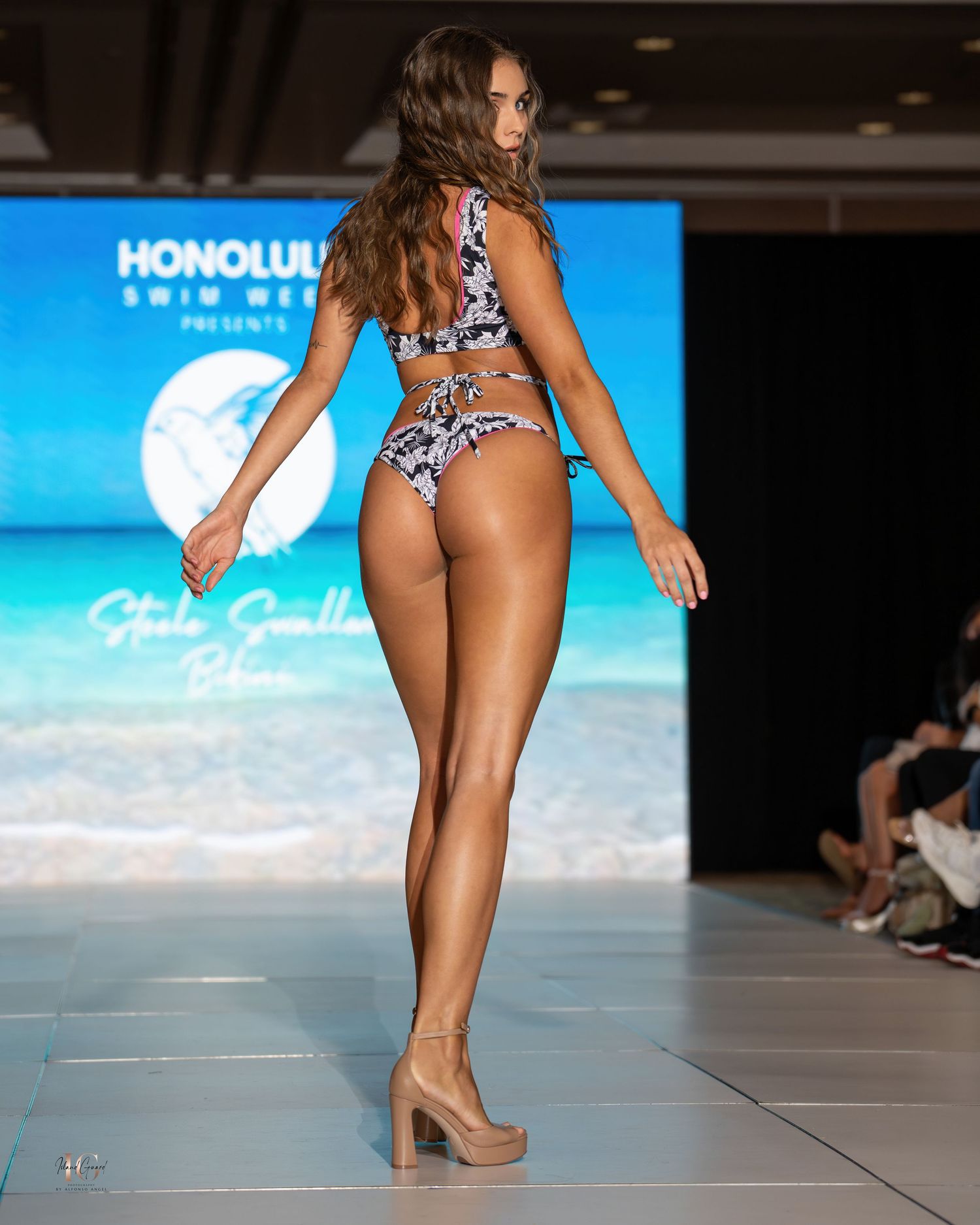 Back view of swimsuit model in patterned bikini walking runway at Honolulu Swim Week fashion show.