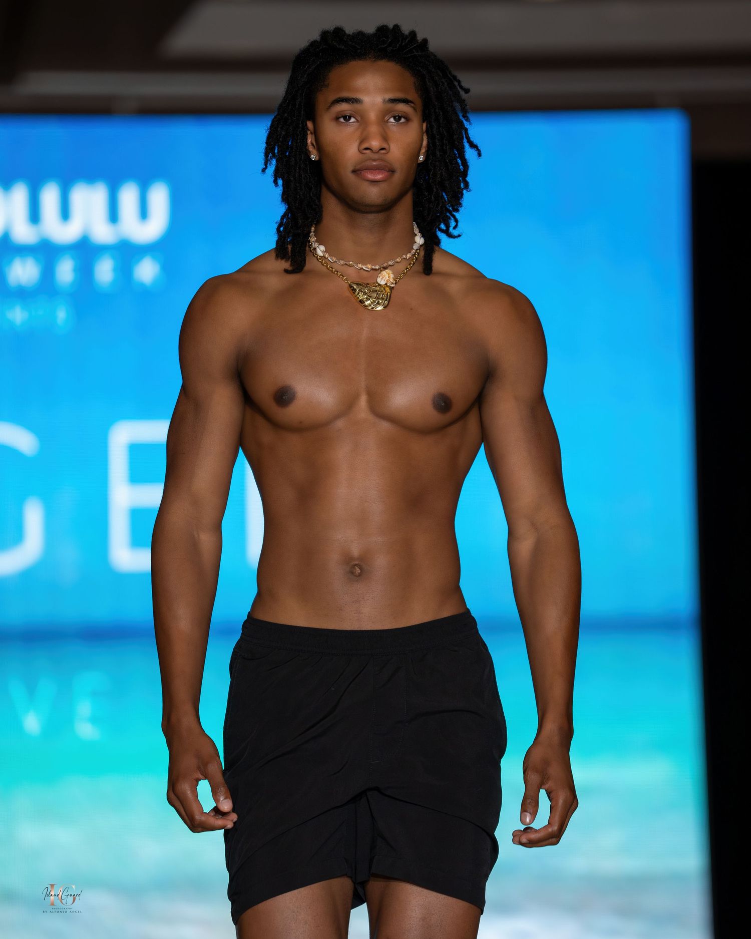 Male model in black shorts walks runway shirtless with gold necklace against blue backdrop.