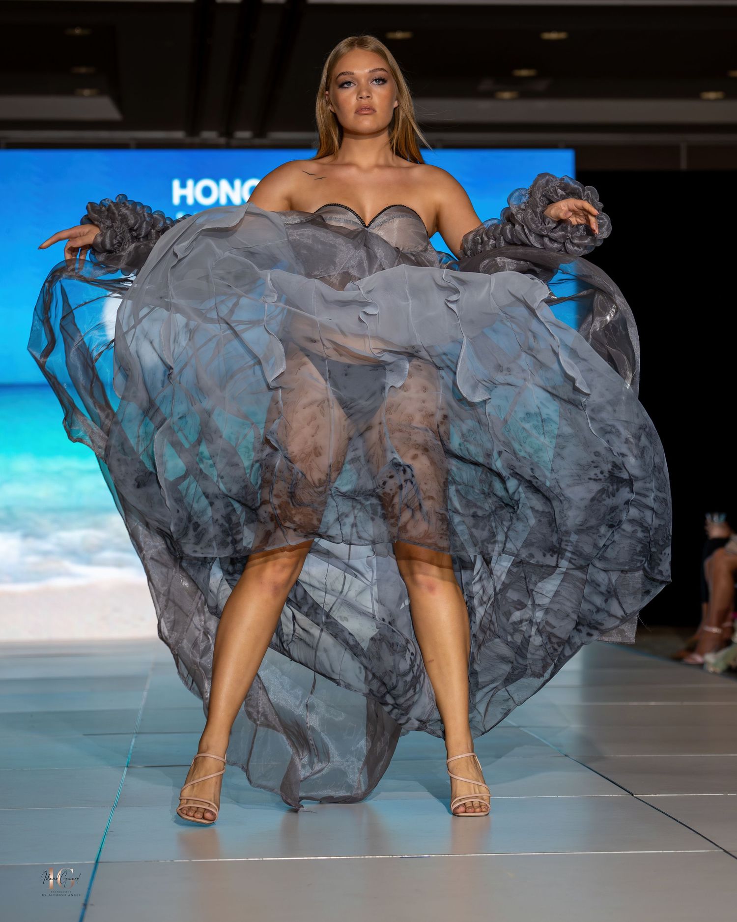 Gray sheer gown with dramatic ruffles and sweetheart neckline showcased on runway with ocean background.