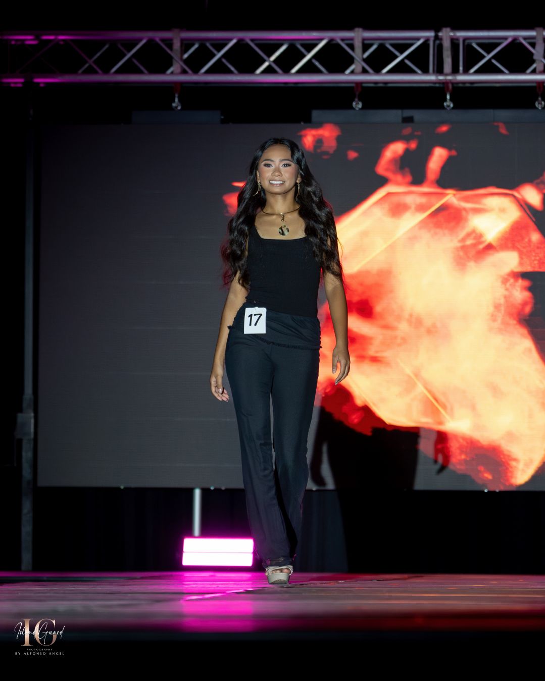 Model walks runway in fitted black ensemble against backdrop of digital flame effects.