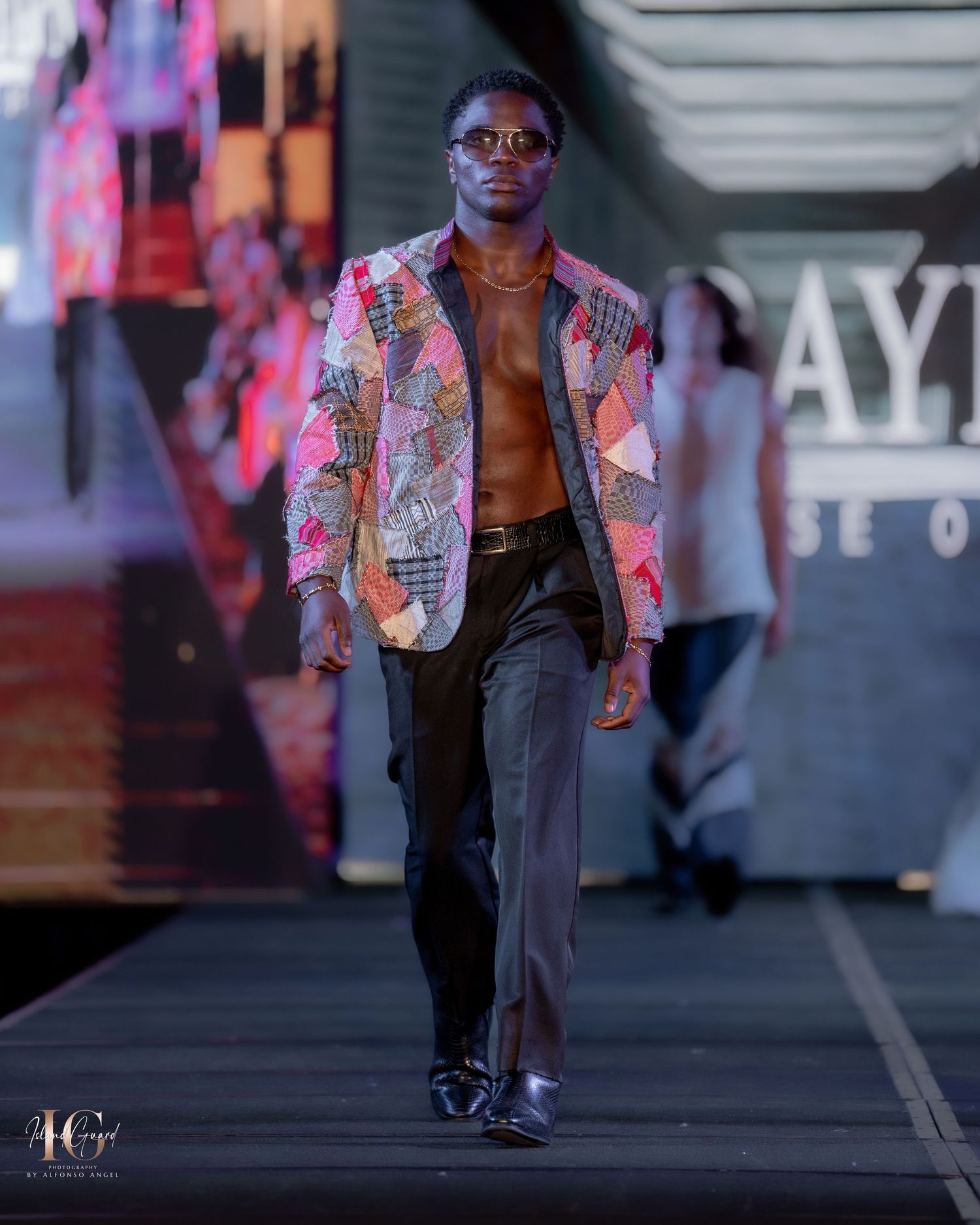 Fashion model walks runway wearing colorful patchwork blazer with sunglasses and dark pants during fashion show.