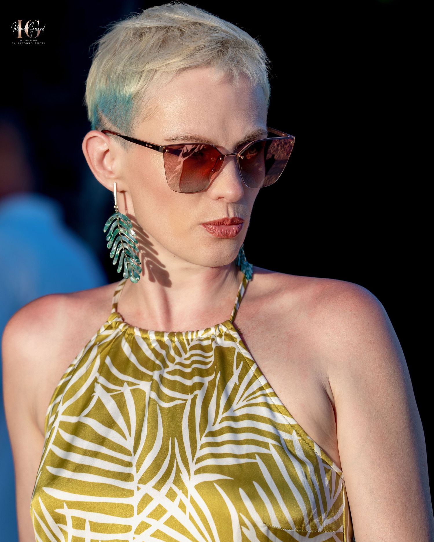 A person with platinum blonde short hair wearing brown sunglasses and a green and white palm print halter dress.