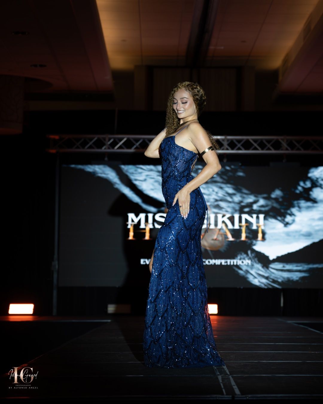 Model in a sparkling navy blue evening gown poses dramatically on stage with event backdrop visible behind her.