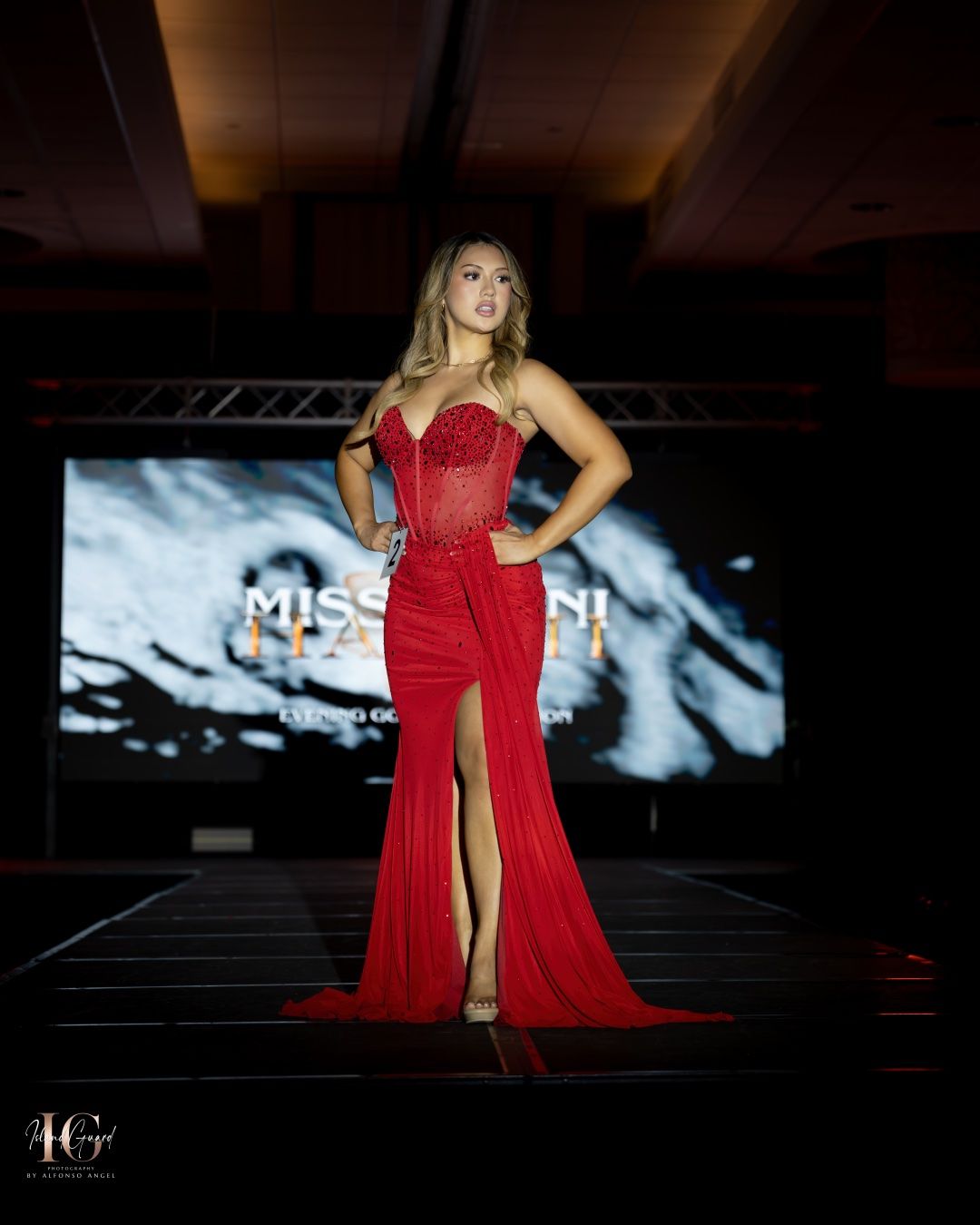 A model walks the runway in a flowing red evening gown with a thigh-high slit and sweetheart neckline on a dark stage.