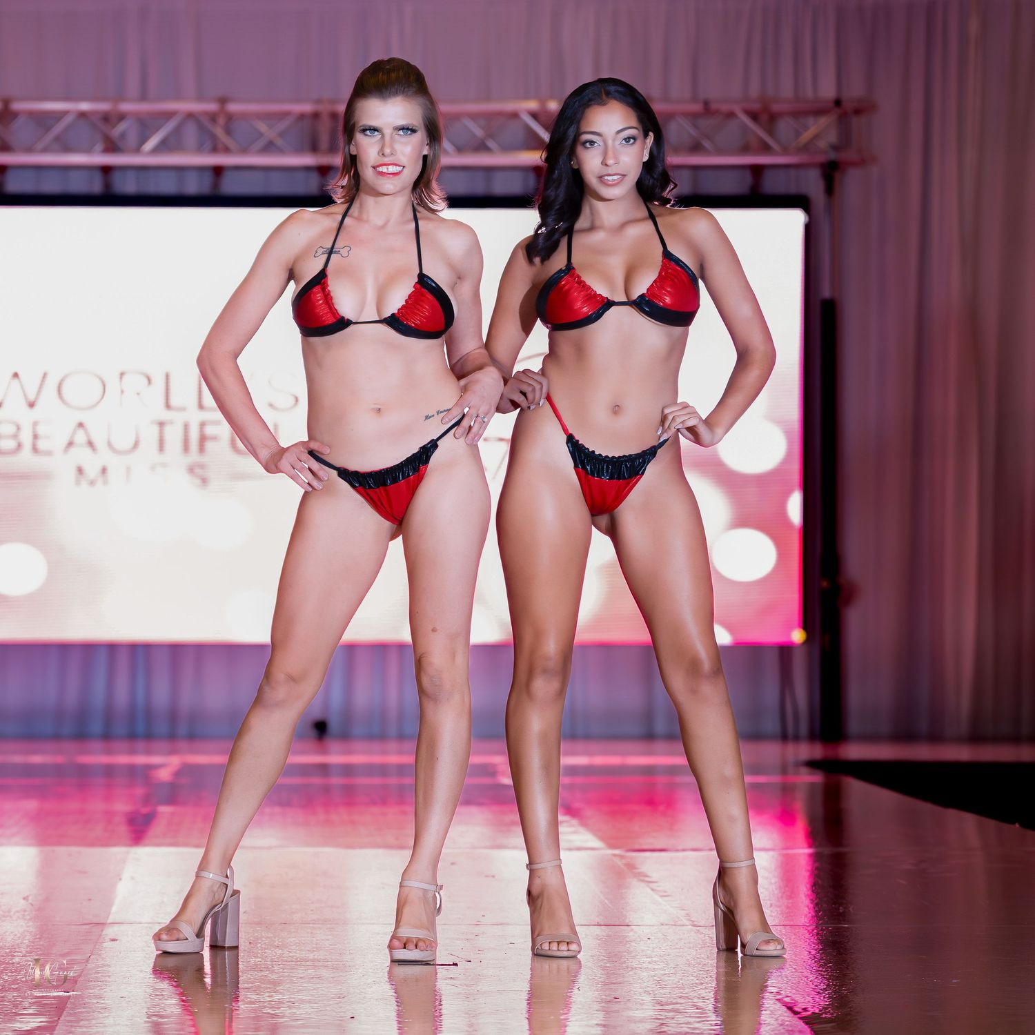 Two models walking runway in matching red bikinis during a fashion show under pink lighting.