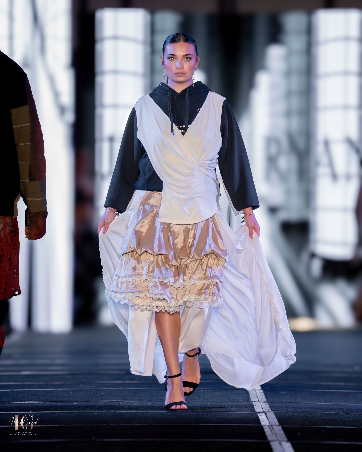 Fashion model walks runway in white avant-garde dress with ruffled details and black sleeves during fashion show.