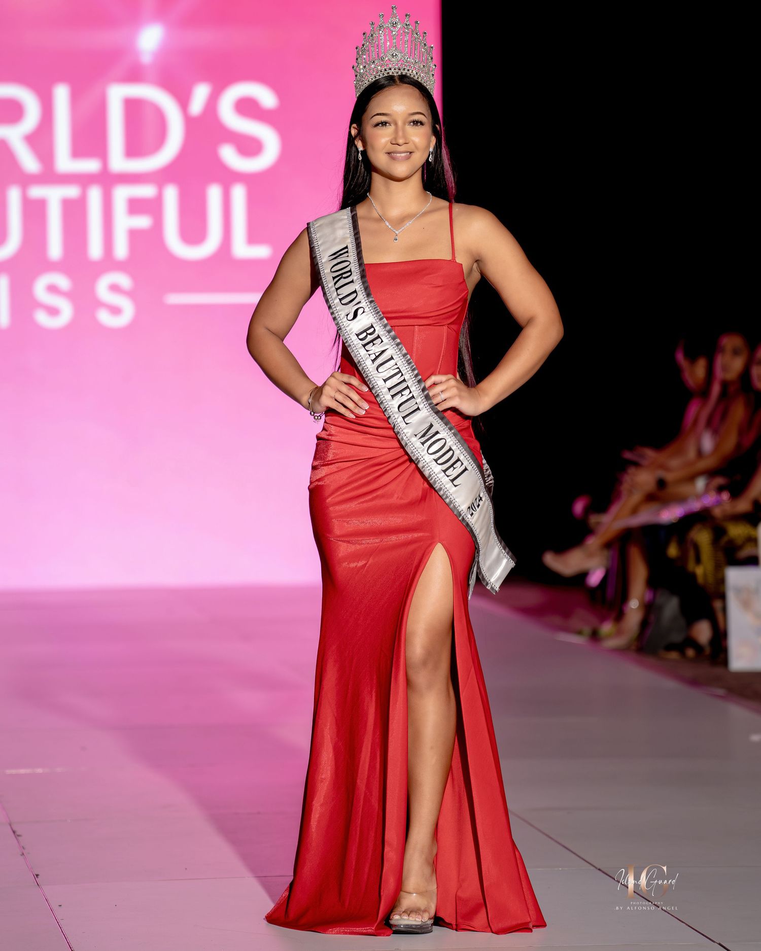 A model walks the runway in a flowing red evening gown with a thigh-high slit at a fashion show event.
