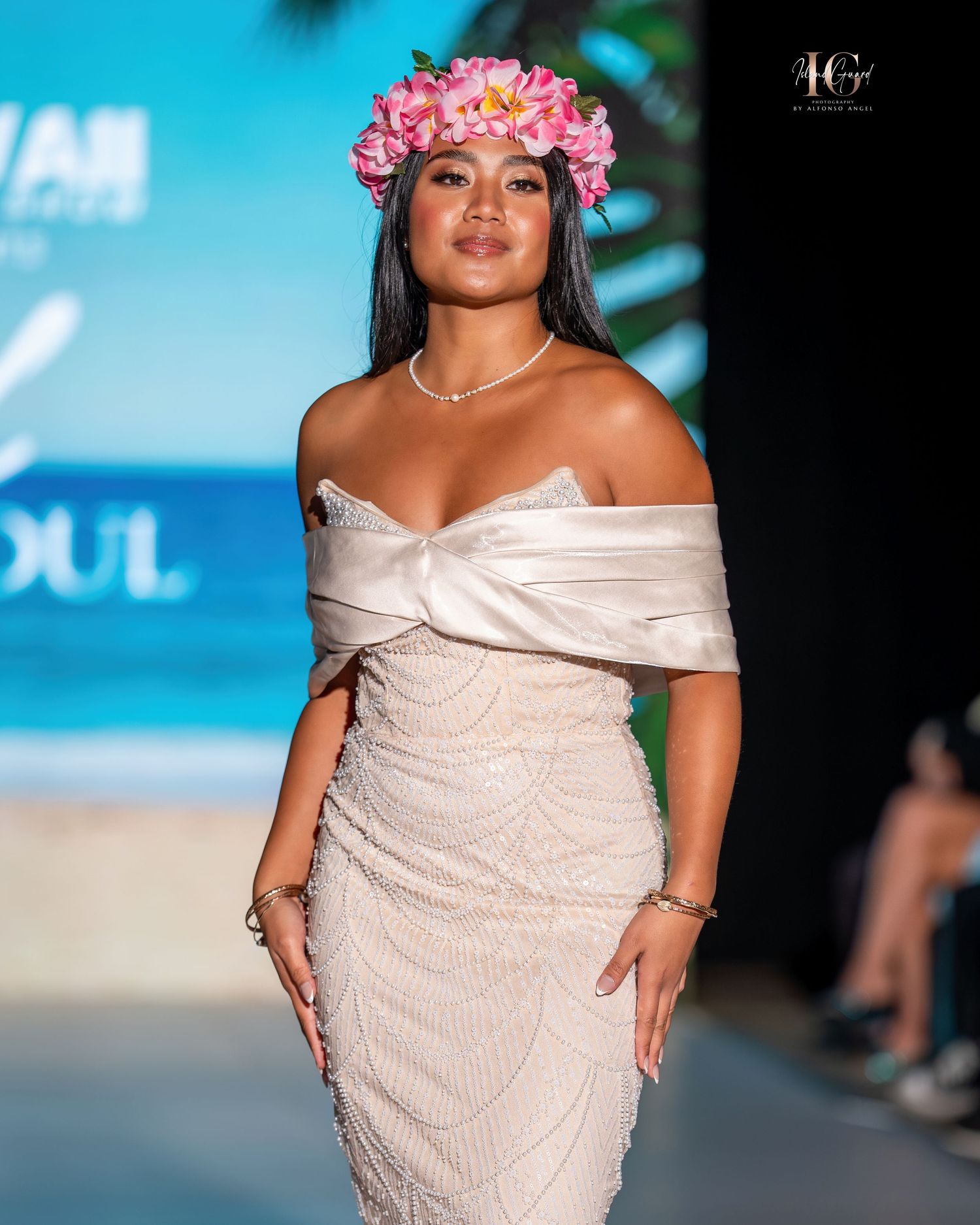 Model walks runway in off-shoulder white dress and pink floral crown during tropical-themed fashion show.