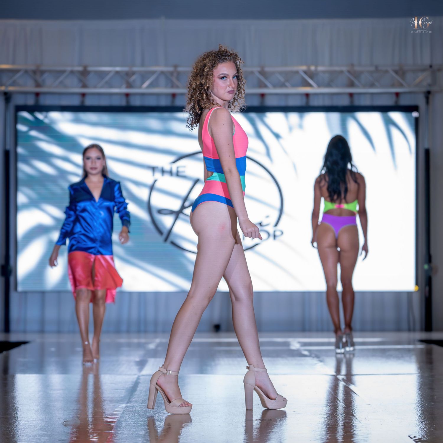 Models walk the runway in colorful swimwear during a fashion show with dramatic lighting and staging.