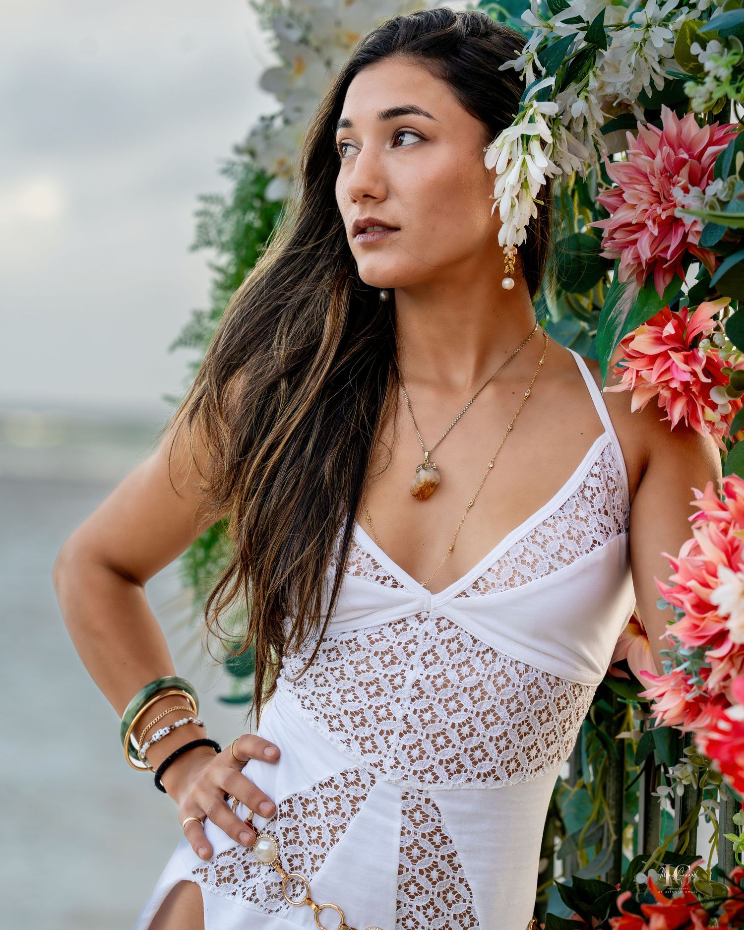 A person in a white lace dress poses against a floral arch with pink blooms and greenery in a tropical setting.