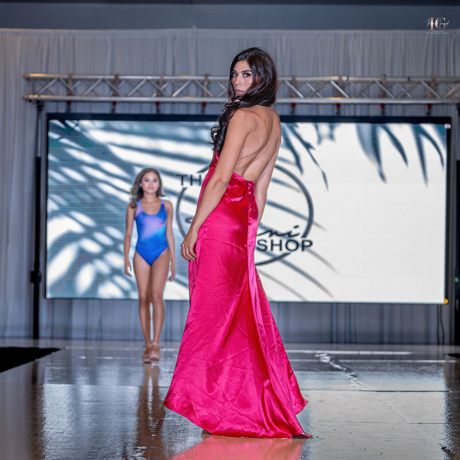 Models walk the runway in vibrant pink and blue dresses during a fashion show on a reflective floor.