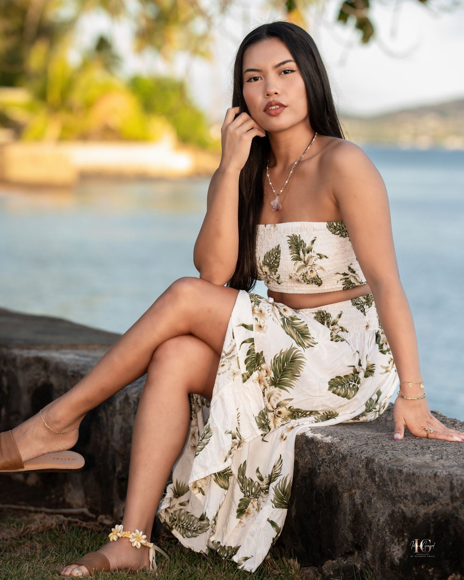 A person in a floral sundress poses elegantly on coastal rocks during golden hour by the water.