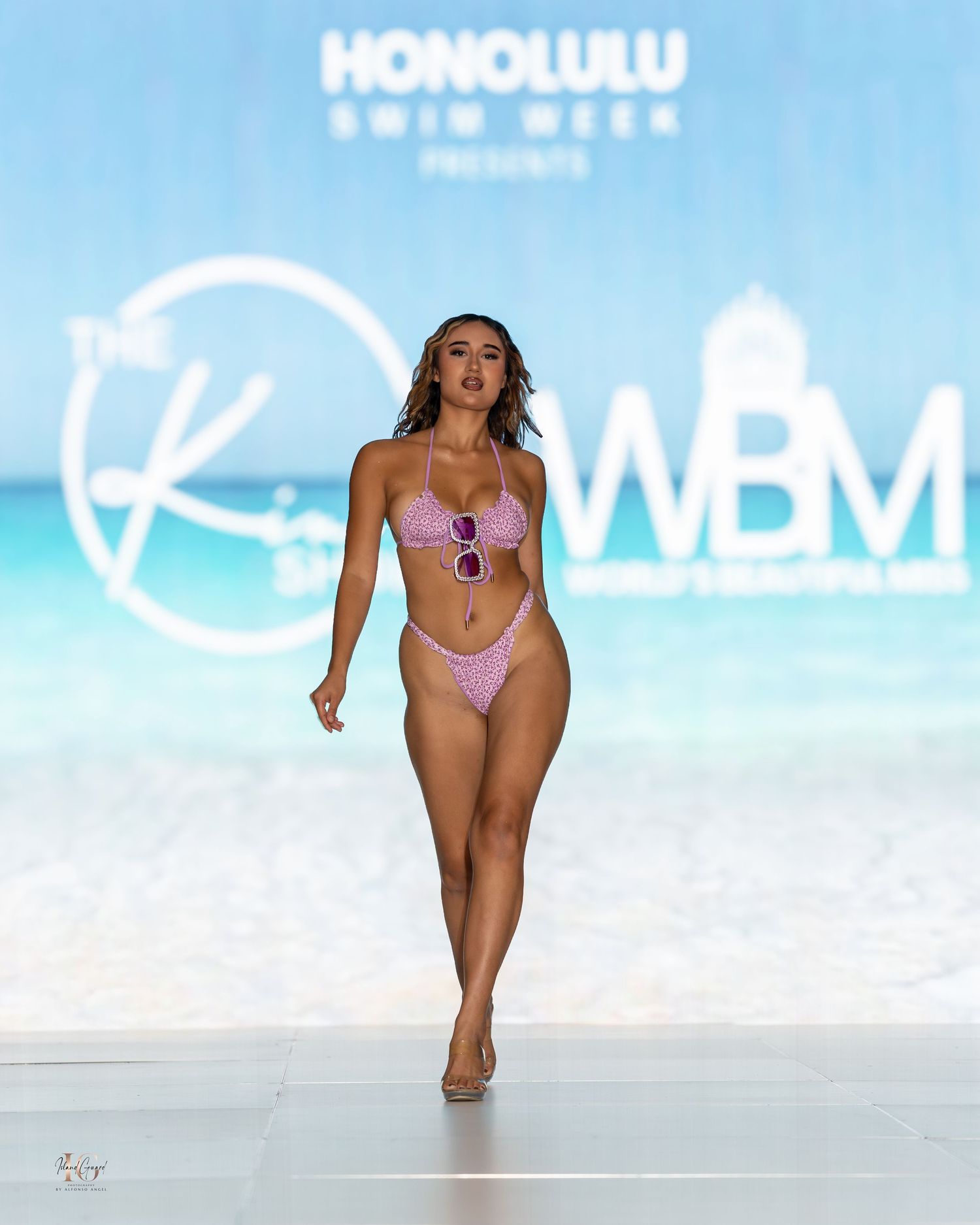 Model showcases purple swimwear on brightly lit runway with Honolulu backdrop.