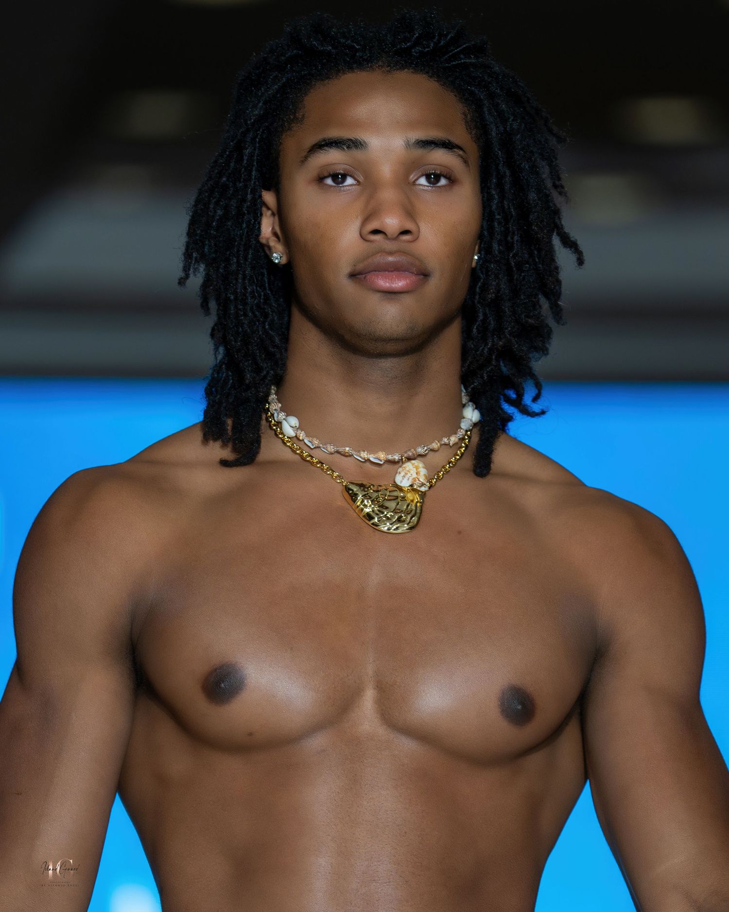 Model with dreadlocks wearing gold chain necklace poses shirtless against blue background.