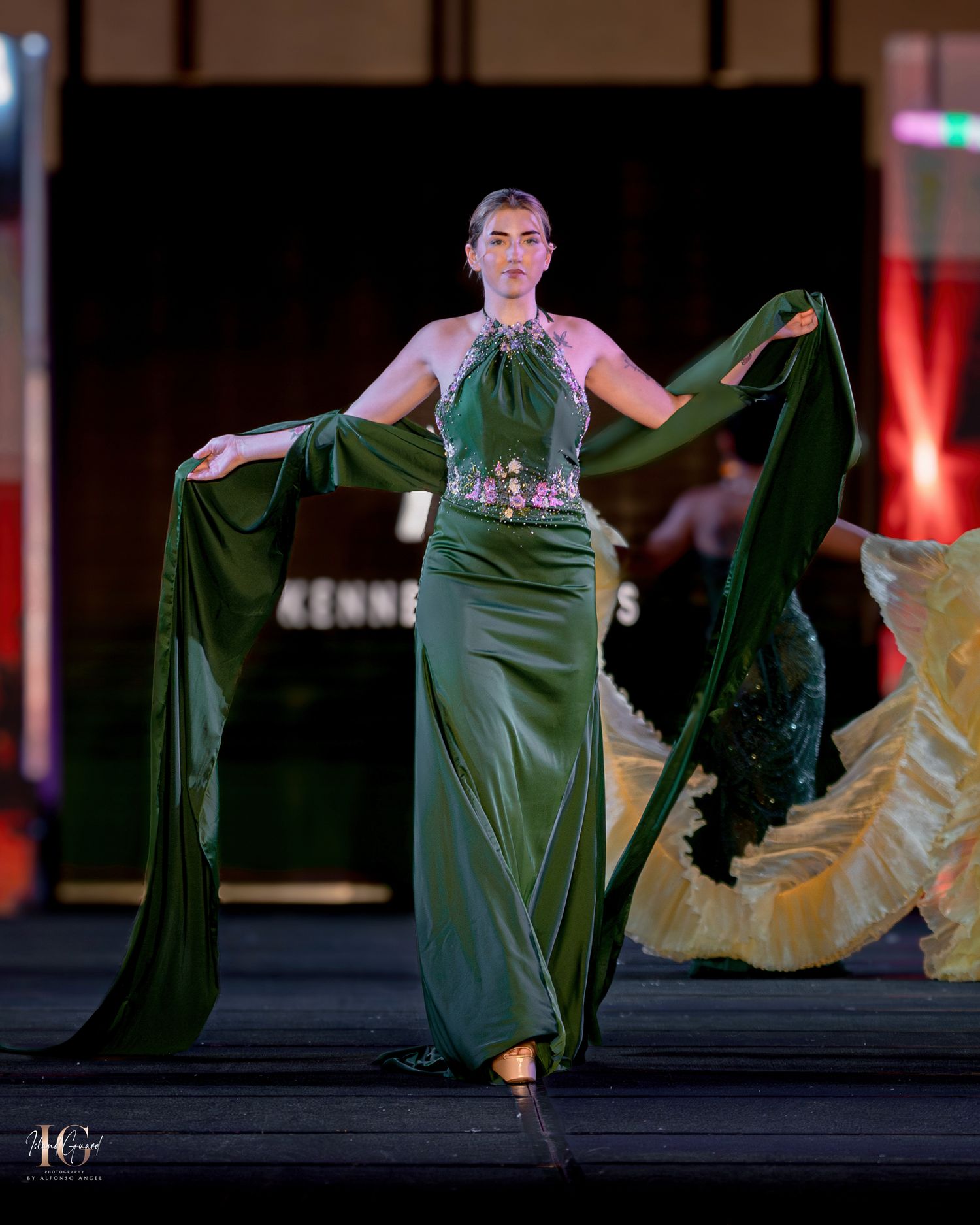 Elegant emerald green evening gown with flowing cape details presented on dark runway.