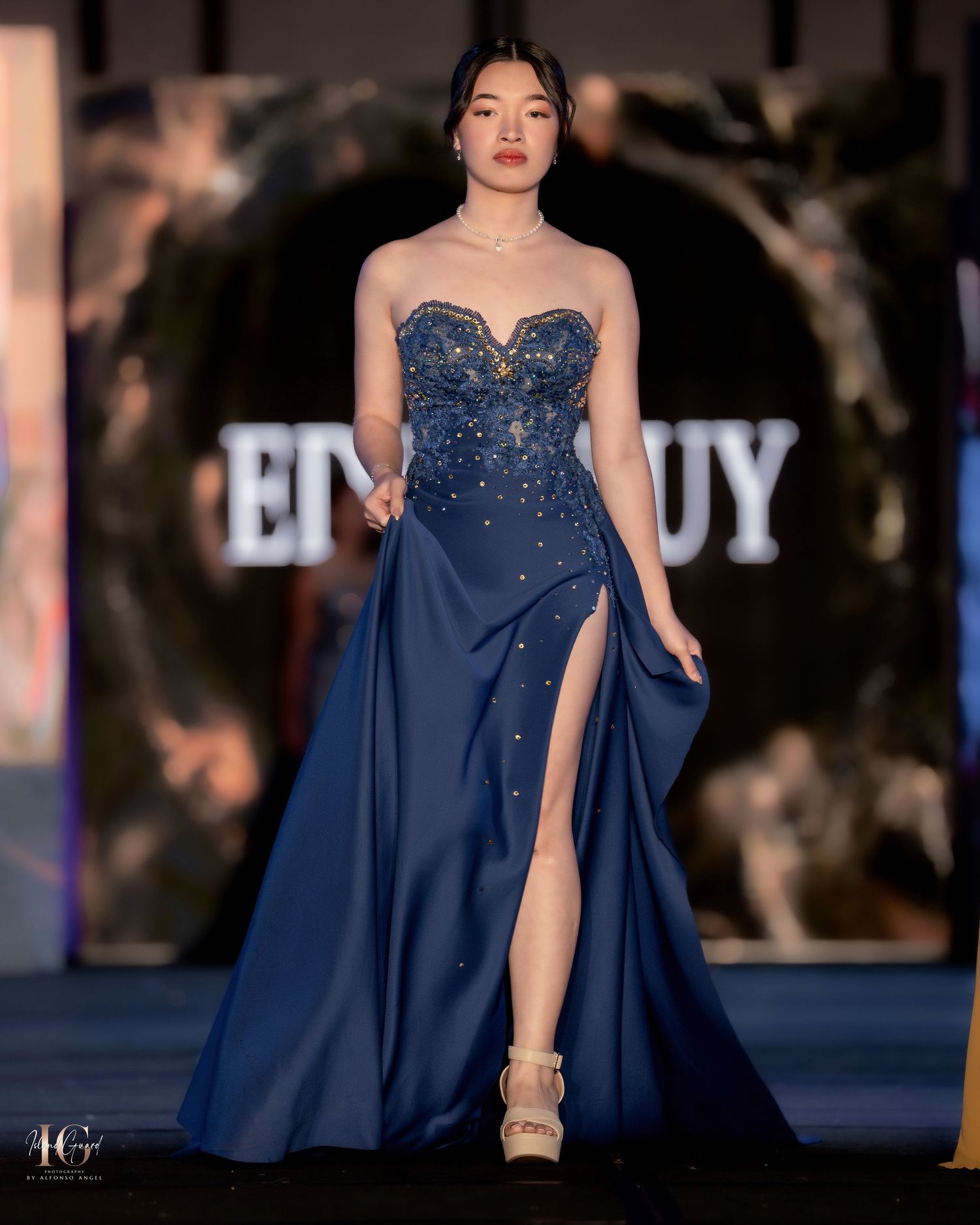 Elegant navy blue evening gown with sparkly embellishments and flowing cape details showcased on fashion runway.