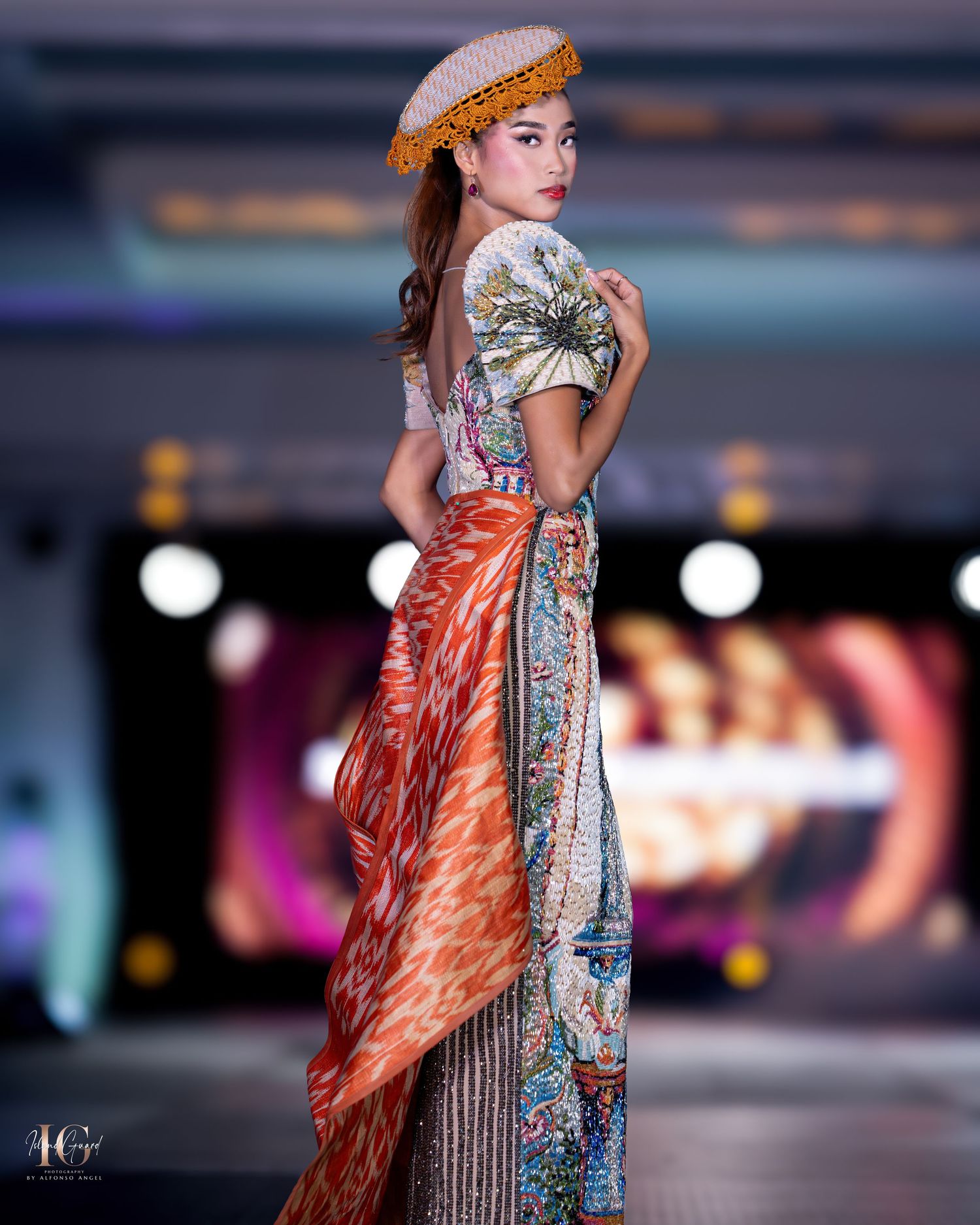Fashion model presents embellished silver gown with orange cape during evening runway show.
