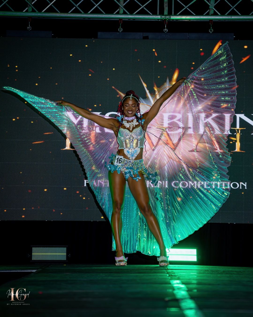 Performer in dazzling iridescent wings poses dramatically on dark stage illuminated by green and blue theatrical lighting.