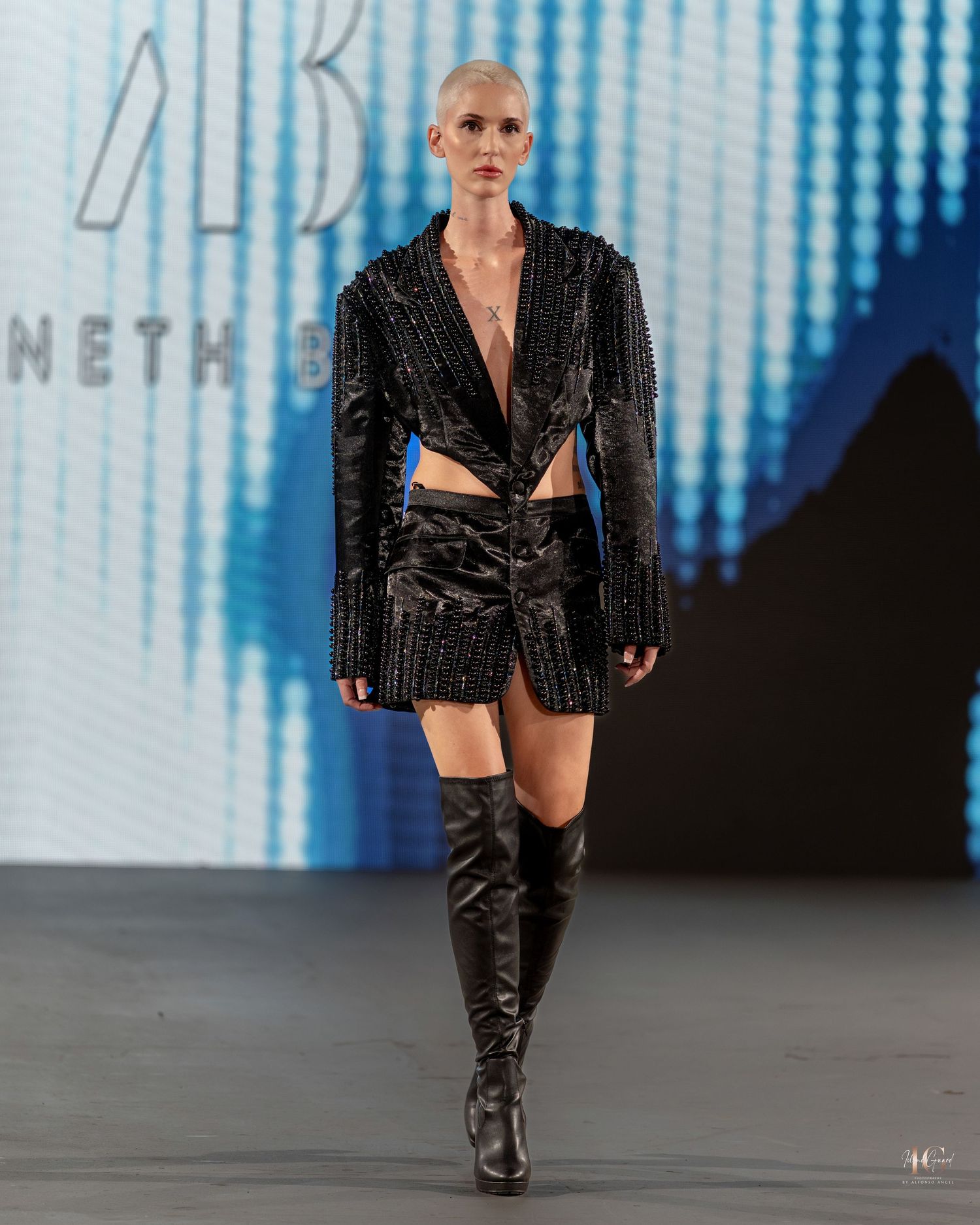 Model walks runway in black sequined blazer and thigh-high boots during fashion show with blue lighting.