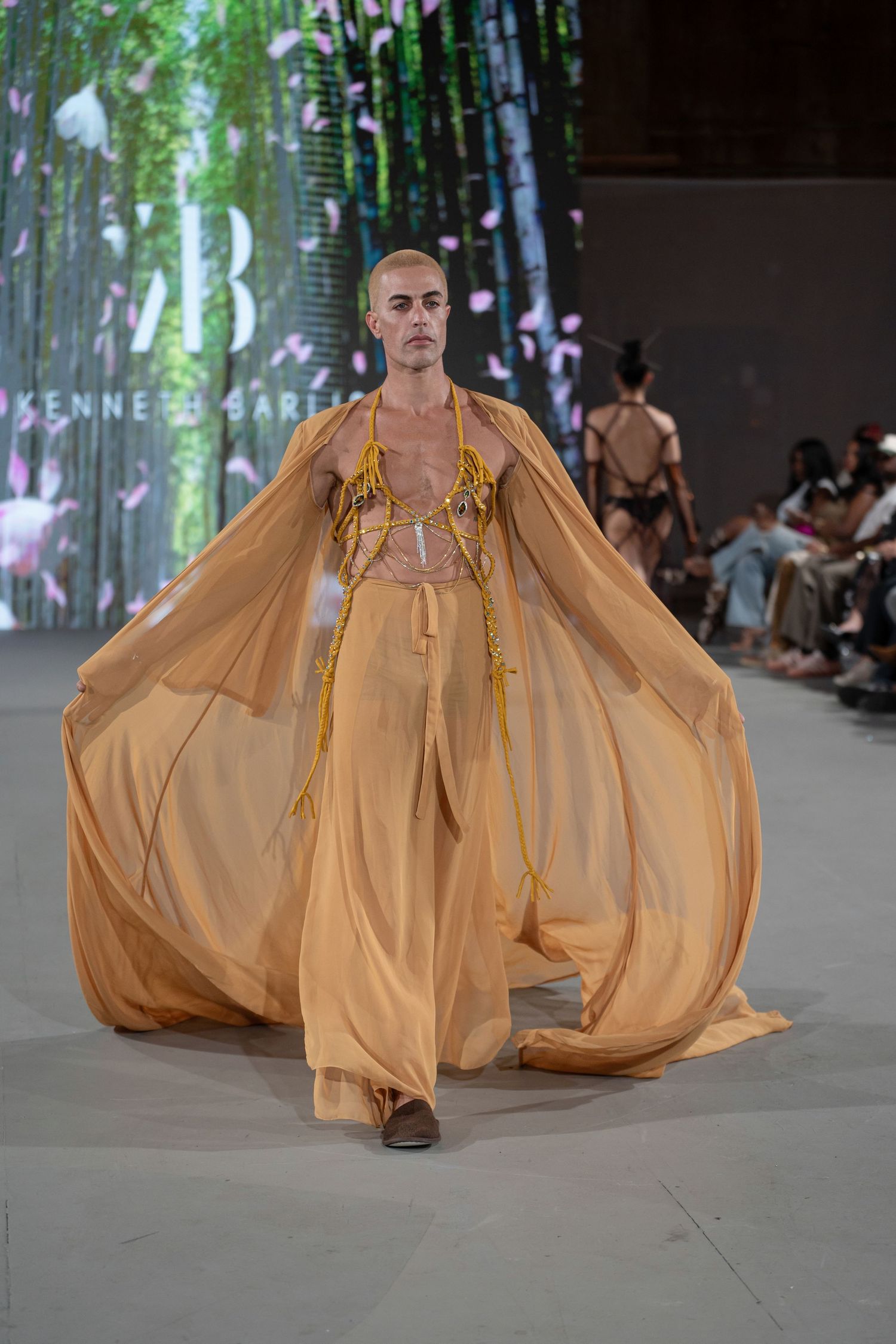 Model walks runway in flowing caramel-colored gown with dramatic billowing silhouette at fashion show.