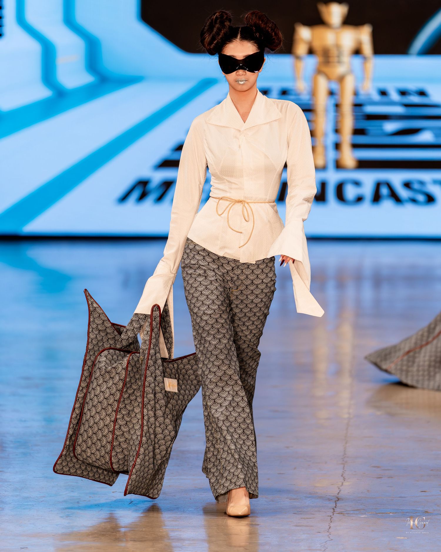Model walks runway wearing white blouse, gray patterned wide-leg pants and designer handbag at fashion show.