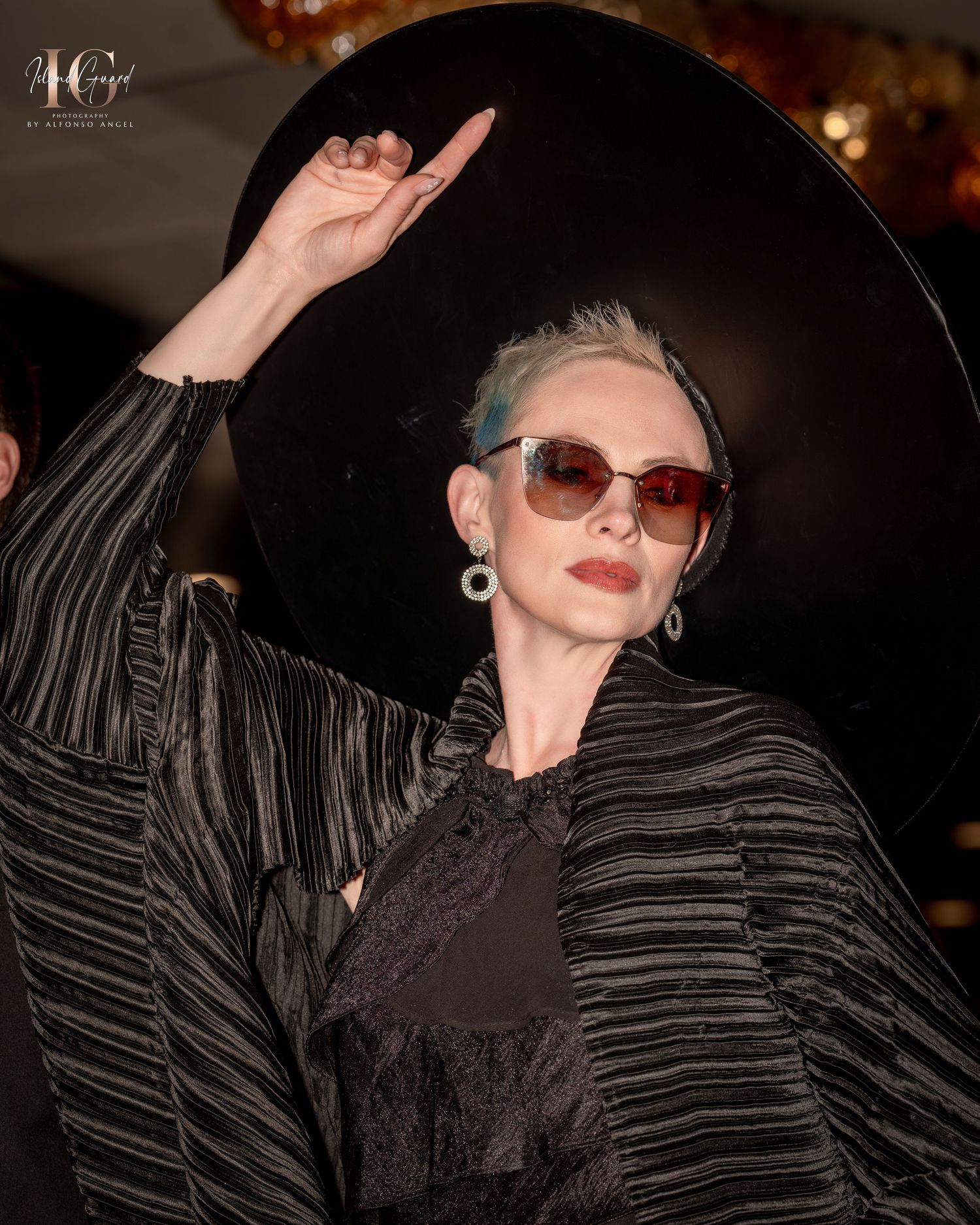 Fashion model in wide brim black hat and striped jacket strikes dramatic pose wearing sunglasses.