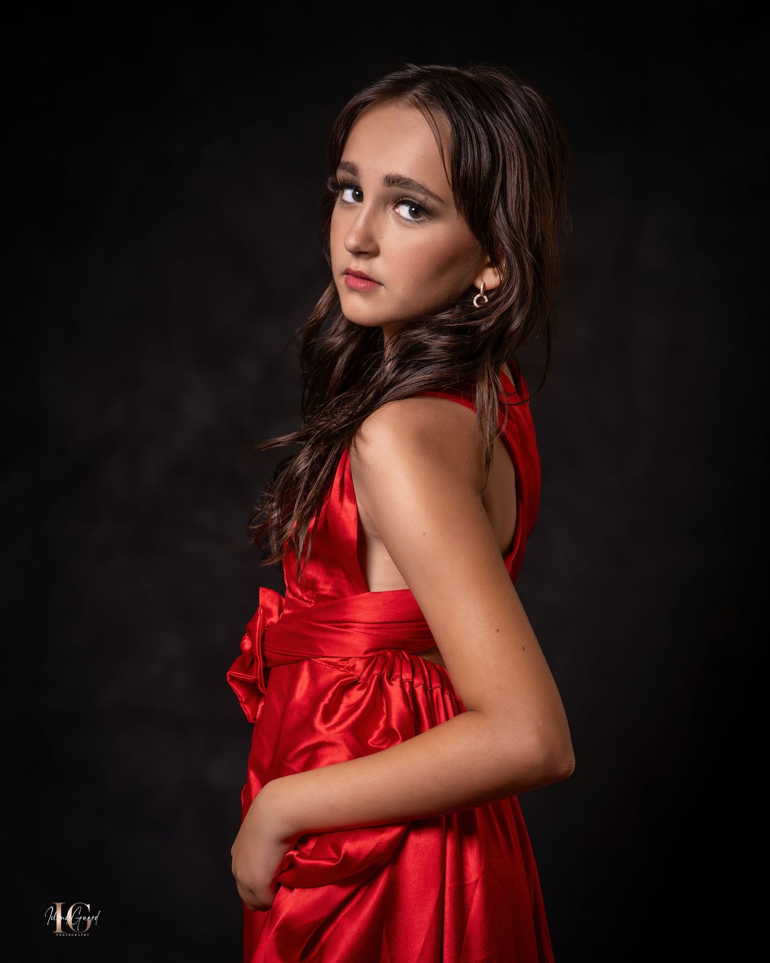 Studio portrait in dramatic lighting shows woman in flowing red satin dress against dark background.